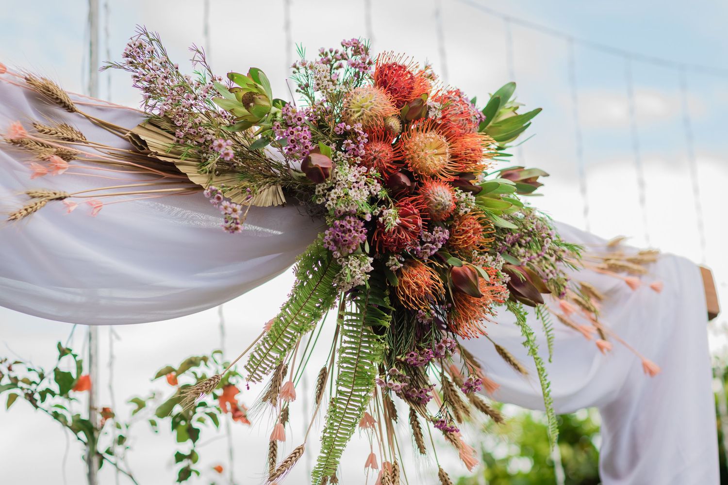 Tropical floral arrangements with protea and greenery decorating a white draped wedding arch.