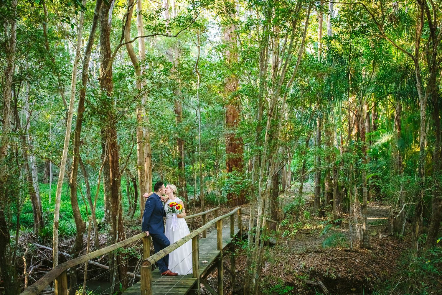 A couple embraces on a wooden boardwalk surrounded by lush green forest at Coolibah downs wedding venue.