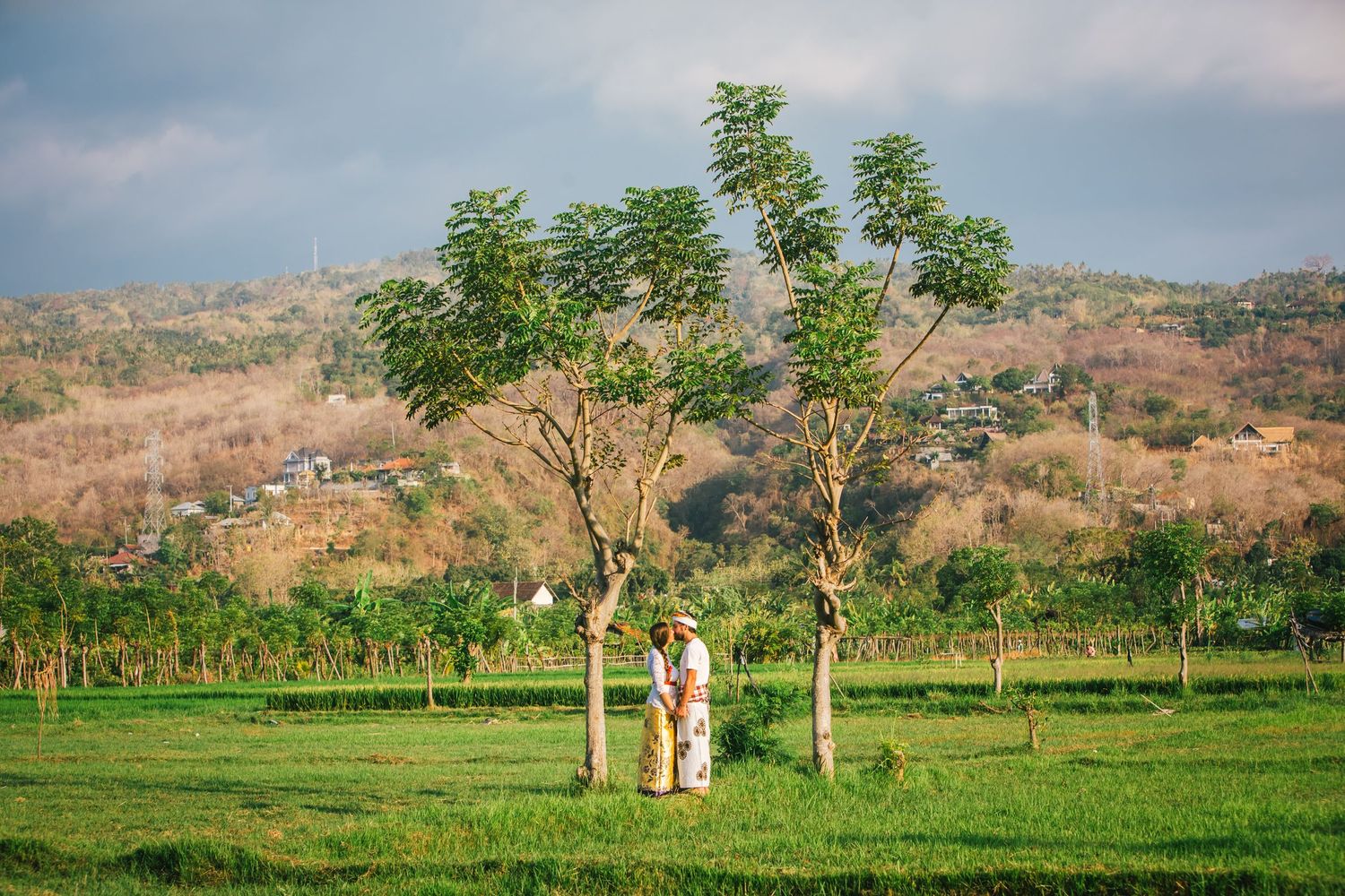 Couple standing beneath trees in sunny green field with hills in background. Bali wedding photogapher