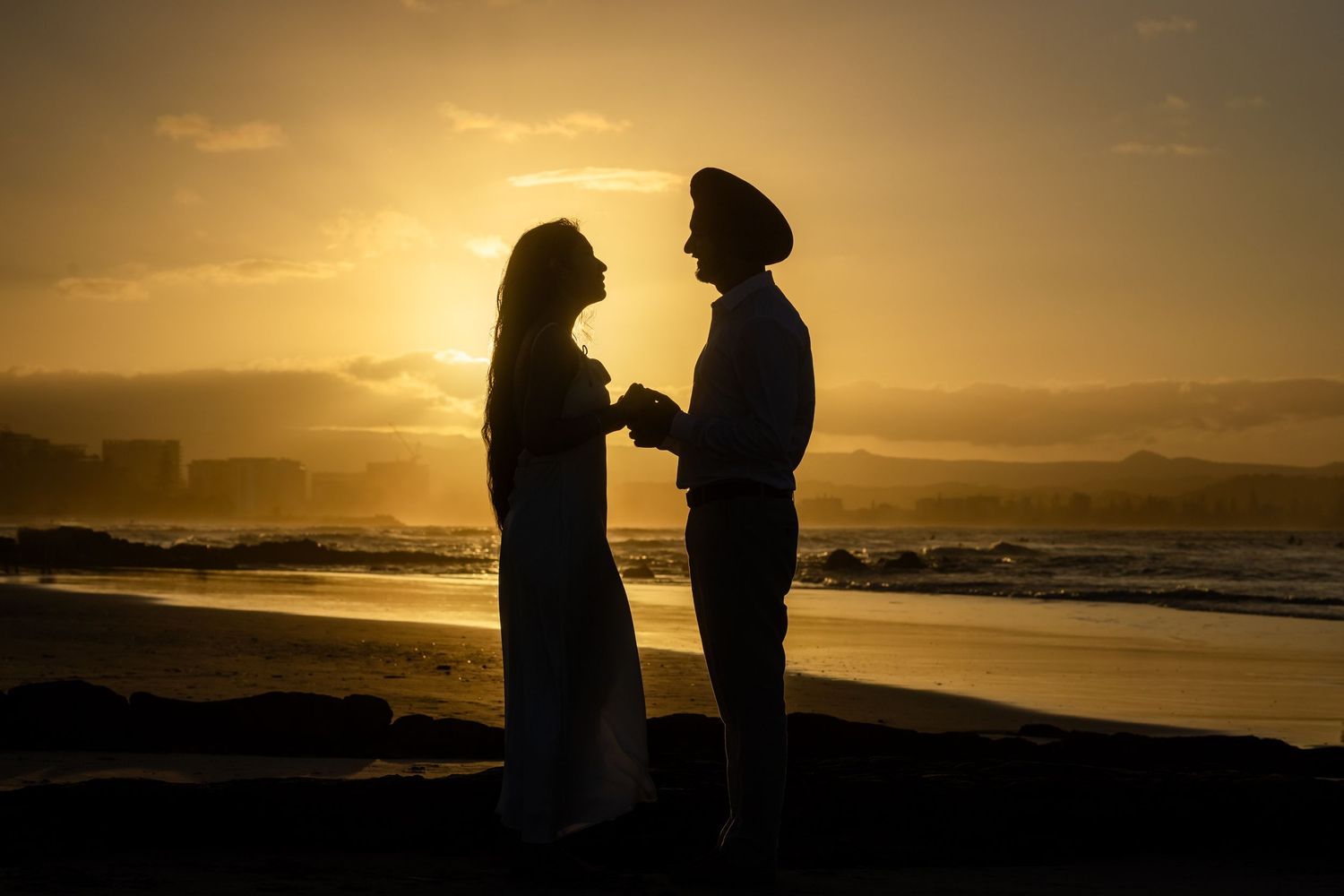 Silhouetted couple embracing on beach at golden sunset with ocean waves in background.