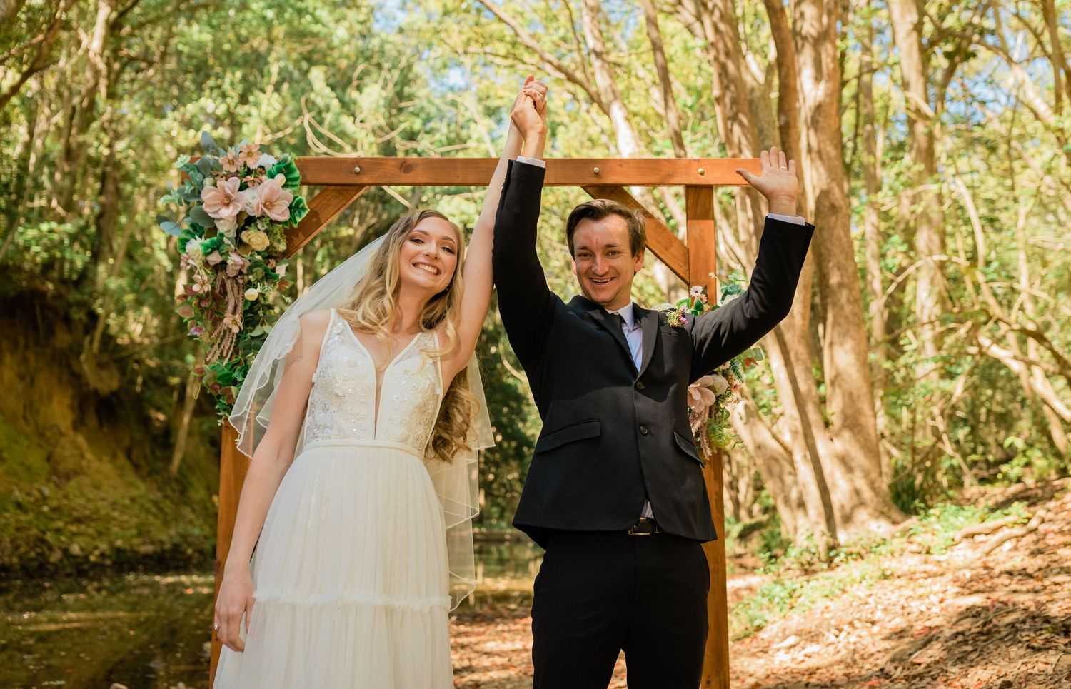 Newlyweds celebrating under wooden arch decorated with flowers in wooded setting.Currumbin Valley wedding photography Gold Coast