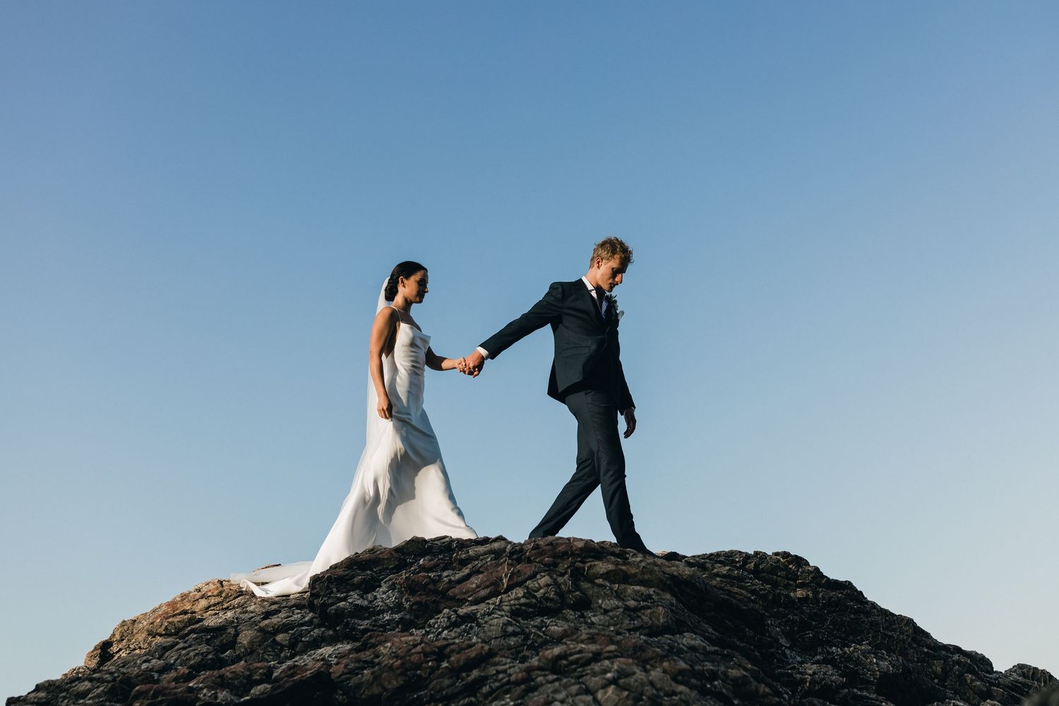 Wedding couple poses on rocky cliff against bright blue sky.