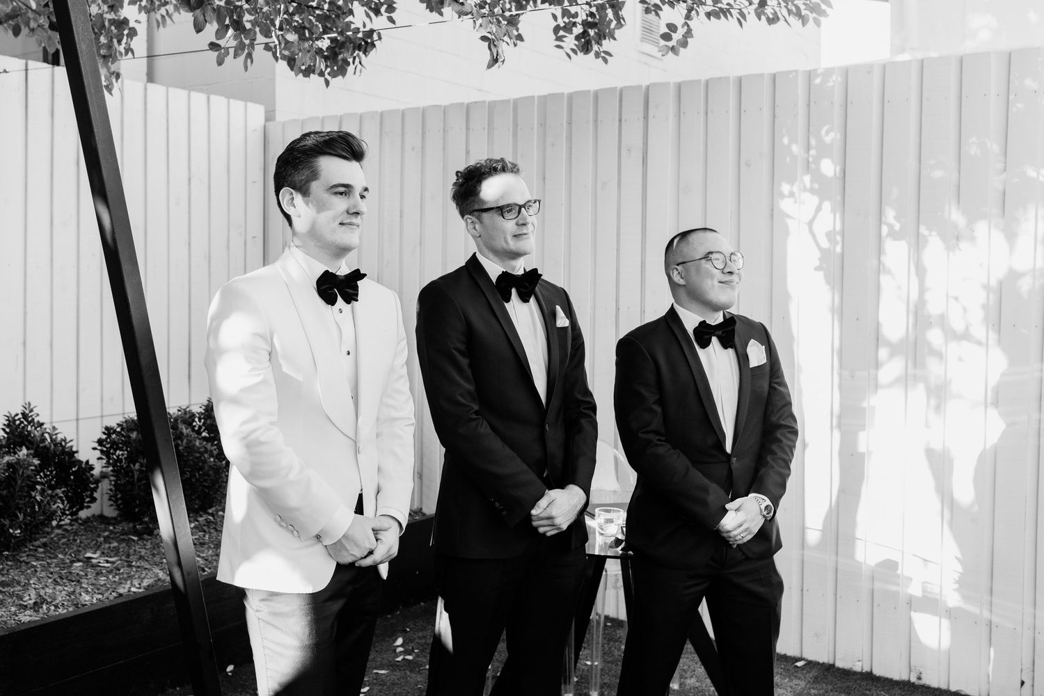 Wedding party gathered in front of white wooden wall outdoors.