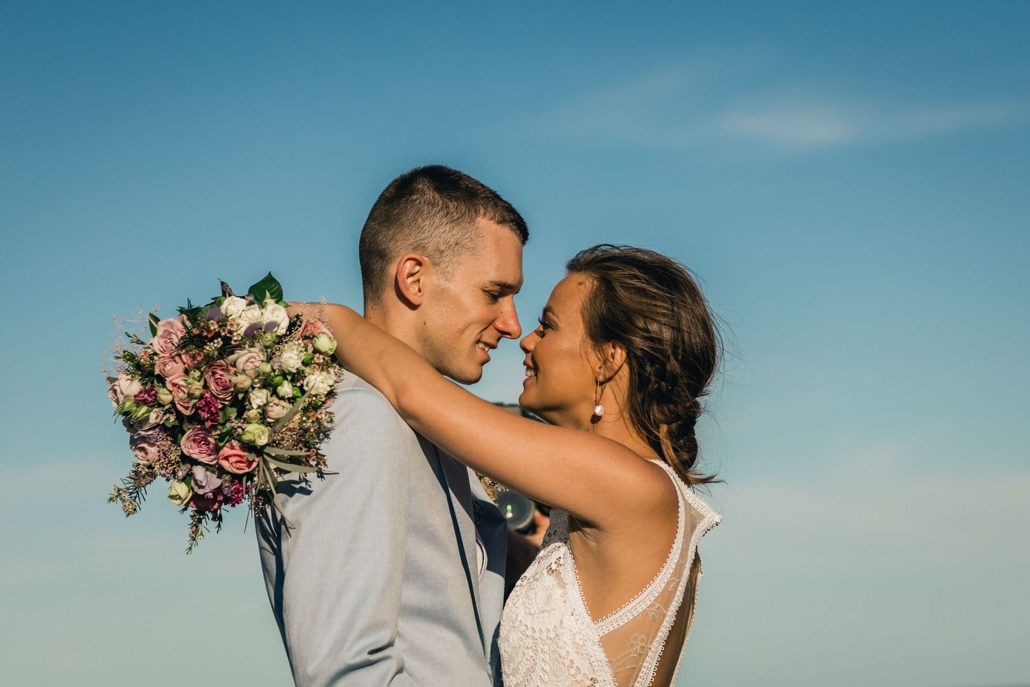 Romantic wedding couple sharing an intimate moment against blue sky with bride holding a pink and white bouquet. Froggies beach wedding