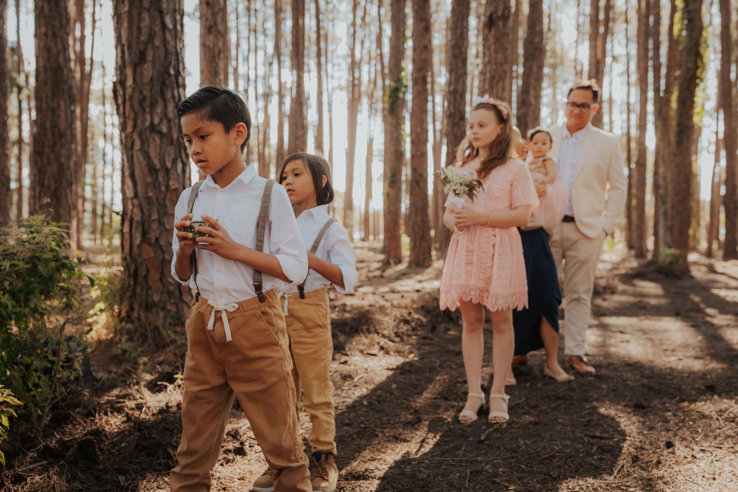 Wedding guests walk along a sunlit forest path during an outdoor ceremony.