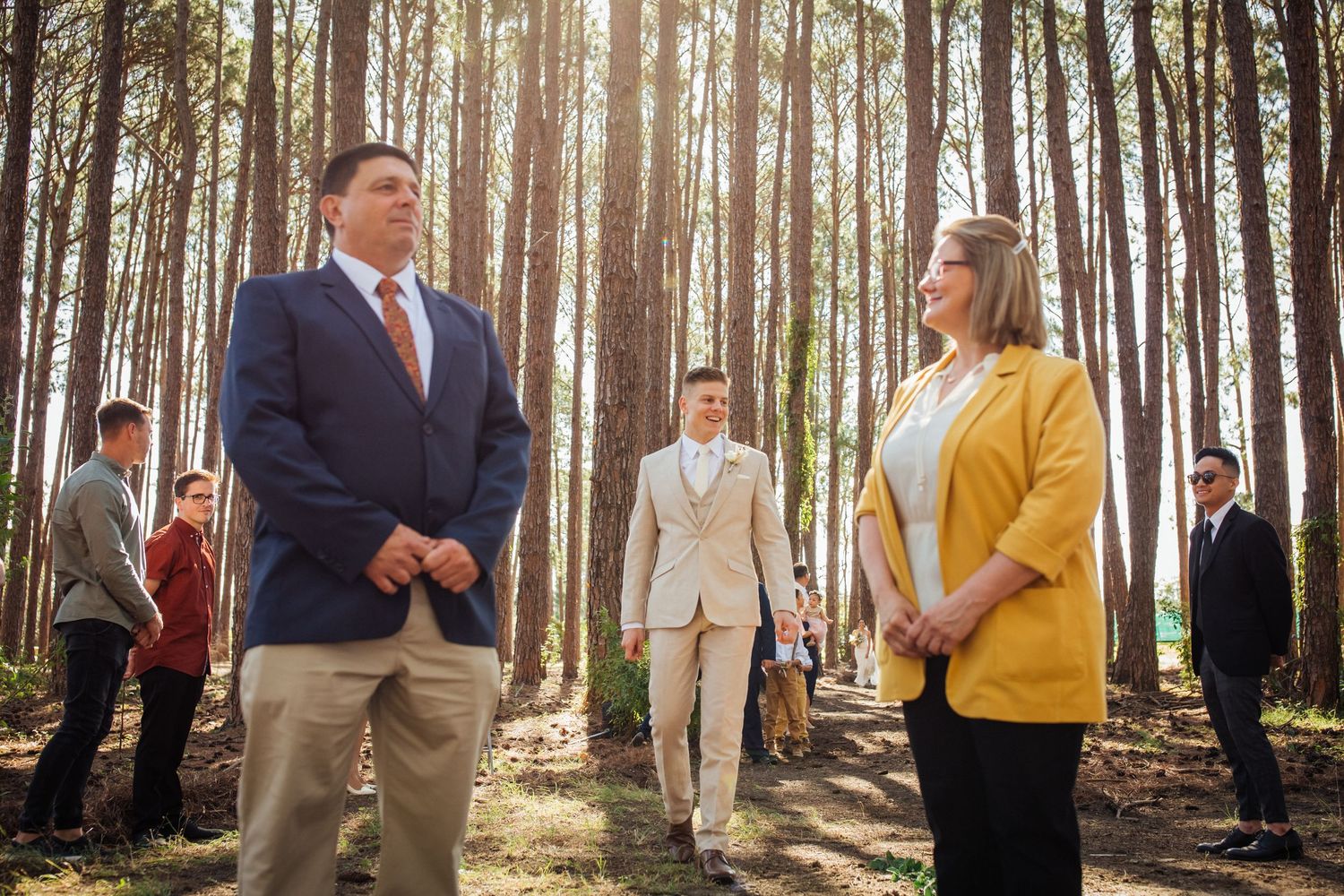 Wedding party members gather in a sunlit forest clearing for formal photographs.