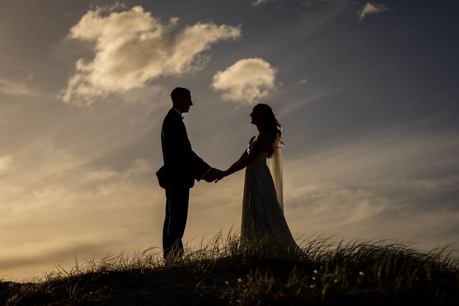 Silhouetted couple holds hands against dramatic sunset sky on grassy hilltop.beach wedding currumbin surf club wedding