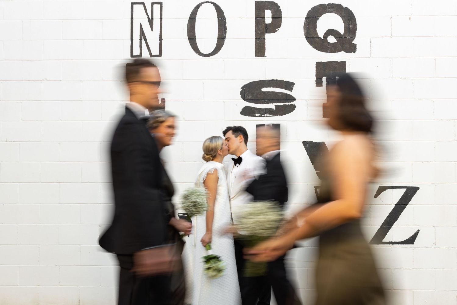 Motion blur of wedding party walking past wall with letters.