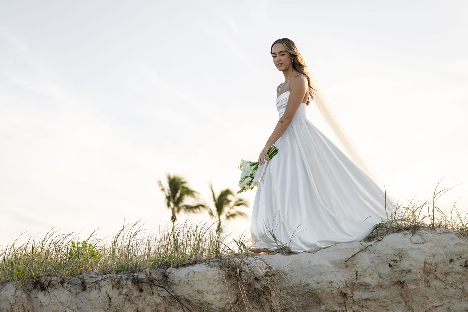 Bride in elegant white dress poses on sand dune with palm trees in background.beach wedding currumbin surf club wedding
