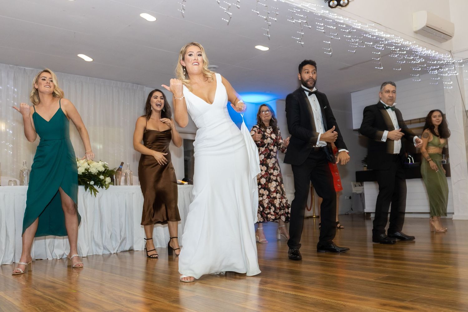 Wedding party dancing in line on wooden dance floor with decorative lights and white decor.
