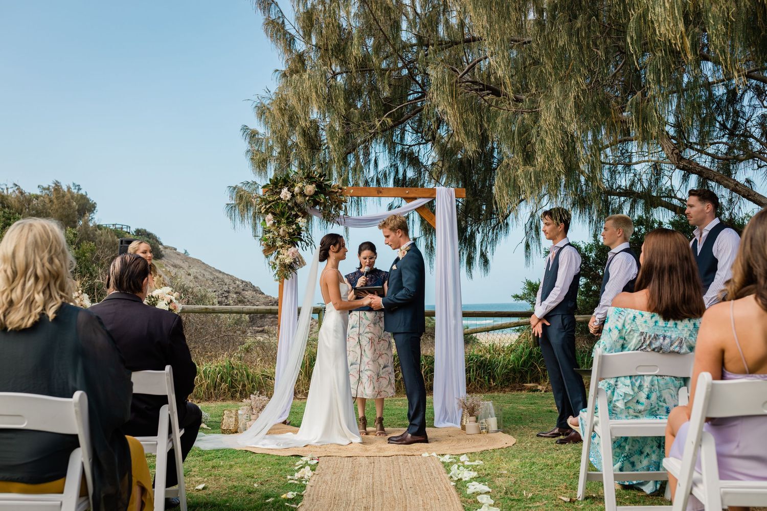 Outdoor wedding ceremony setup with white chairs and flowing fabric under willow trees.