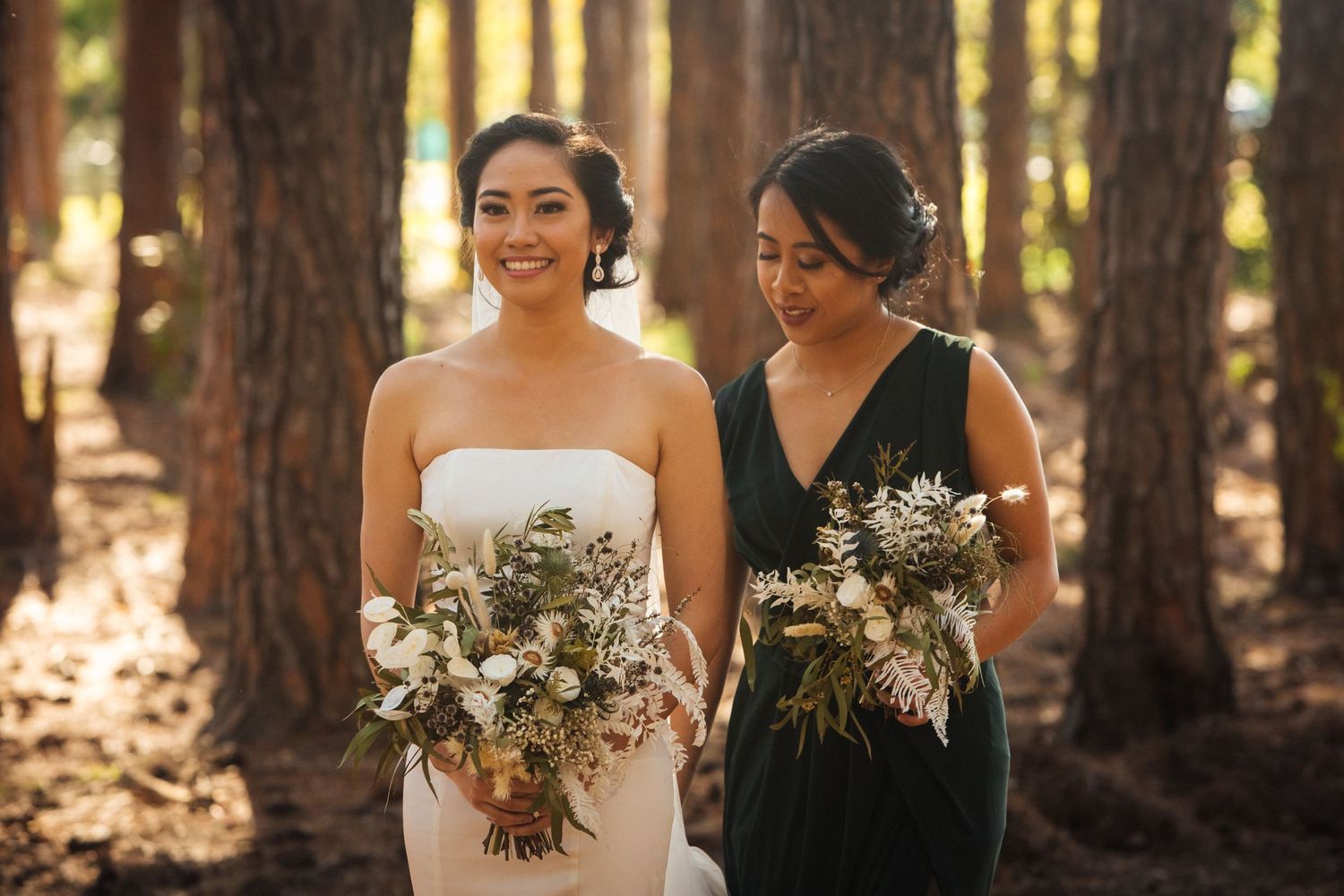Two attendants share a joyful moment while holding bouquets in a forest setting.