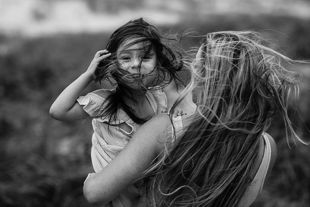 Black and white photo of two girls with wind-blown hair embracing outdoors in a candid moment.
