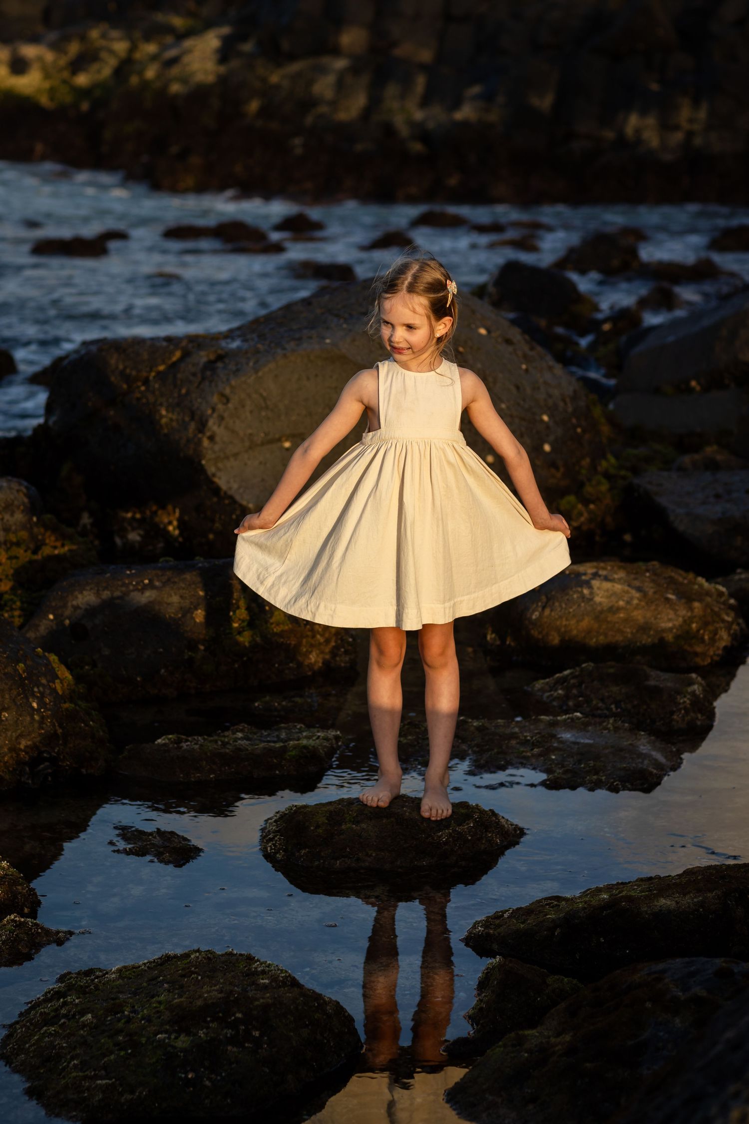 Figure in a cream dress twirls on coastal rocks at sunset with ocean waves in background. Gold Coast candid family photographer.