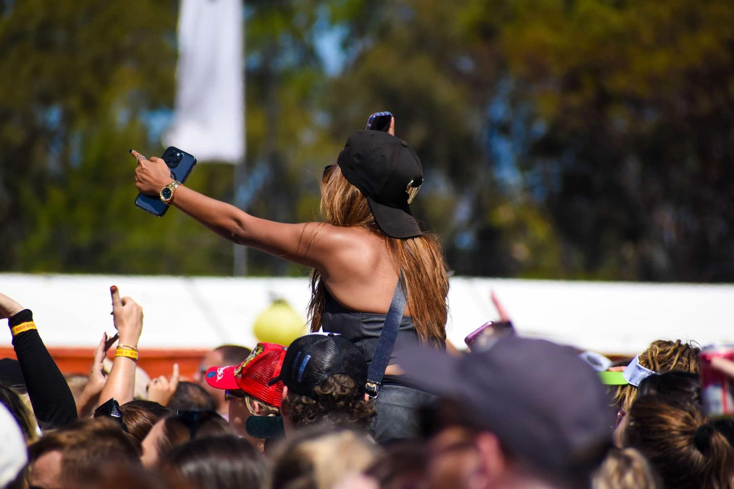 A concert attendee sits on the shoulders of someone above the crowd at an outdoor music festival, holding up a phone.
