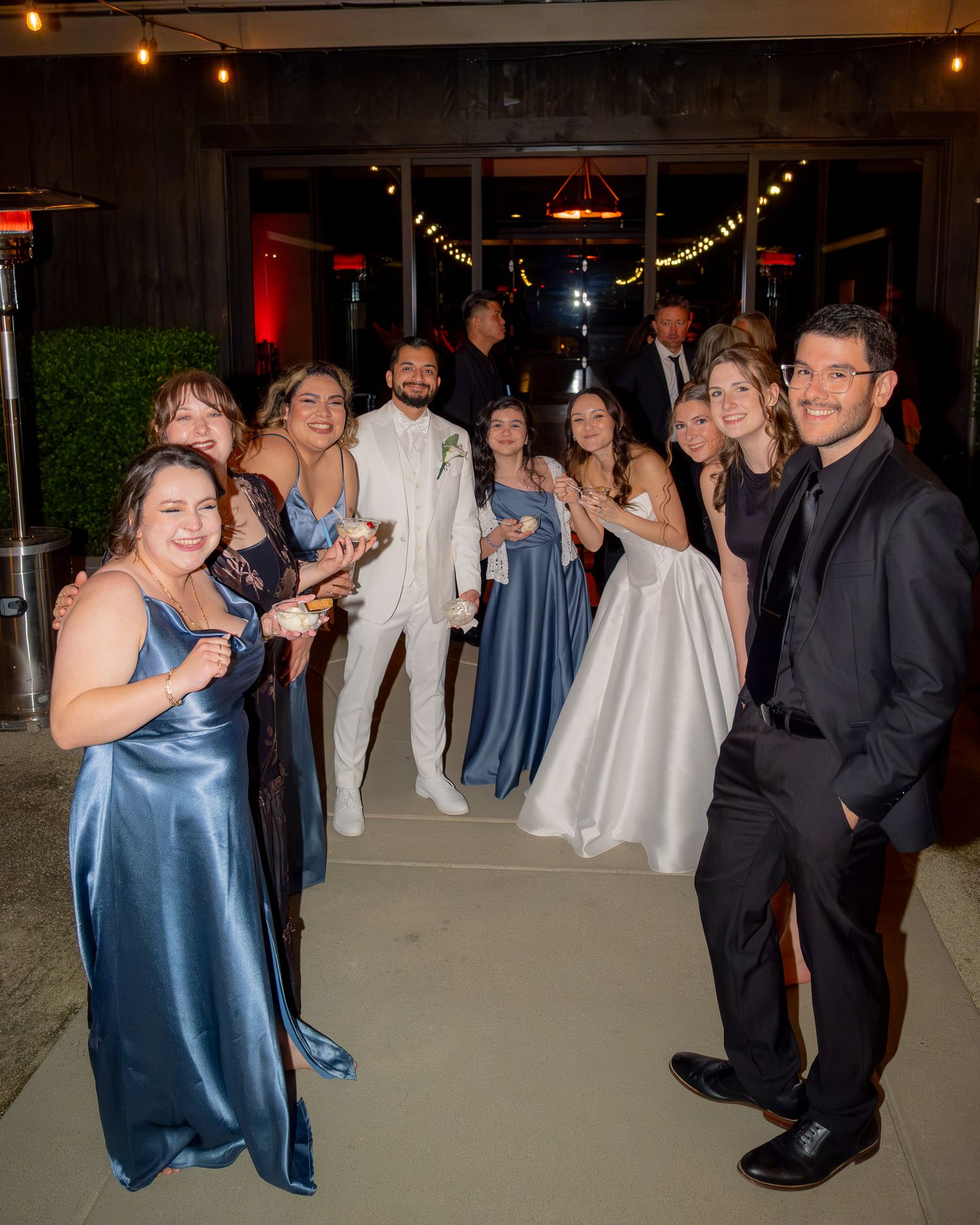 Wedding party group photo outdoors at night, bridesmaids in blue satin gowns with bride and groom in white formal attire.