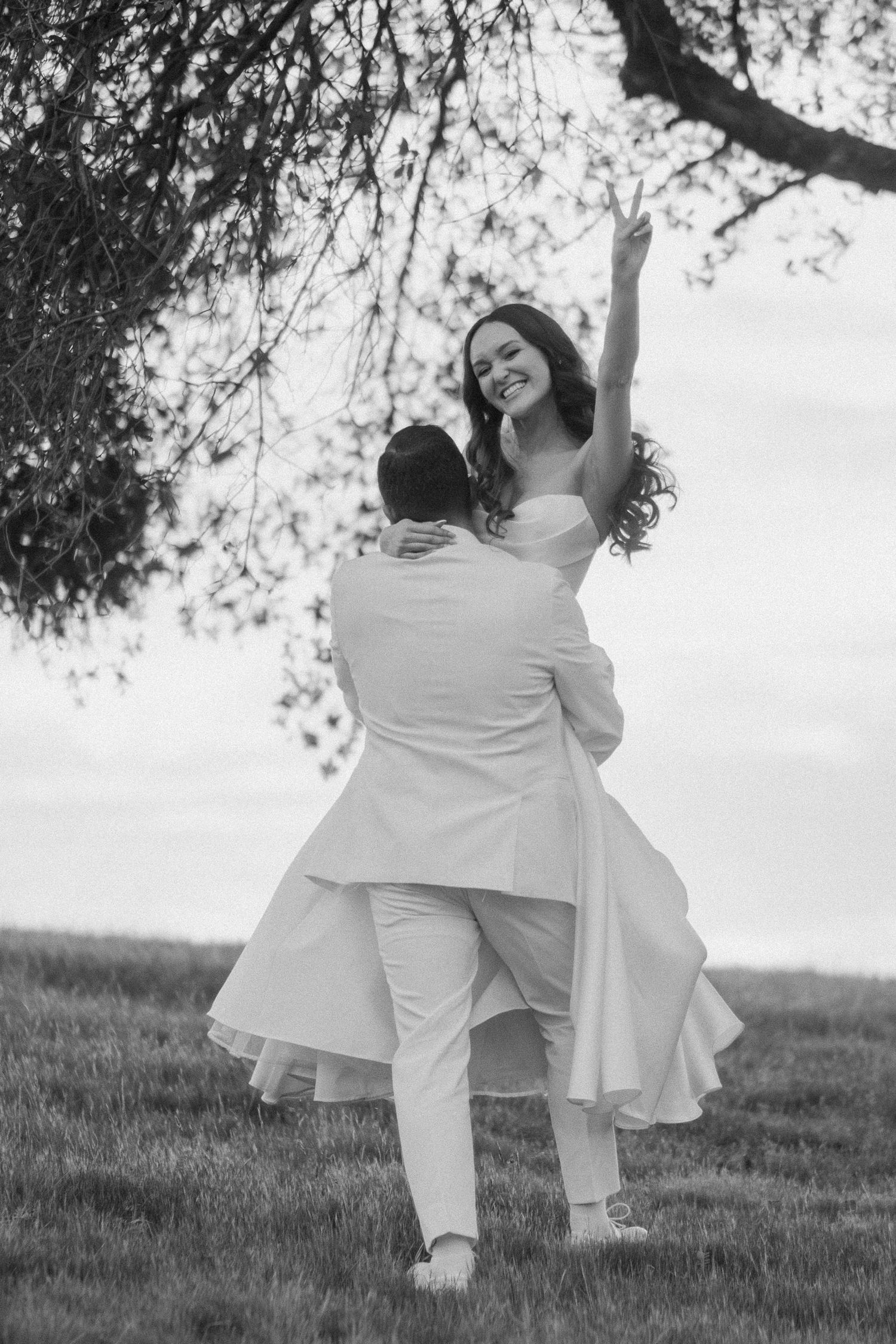 Groom lifting joyful bride in white dress outdoors under a tree in black and white photo.