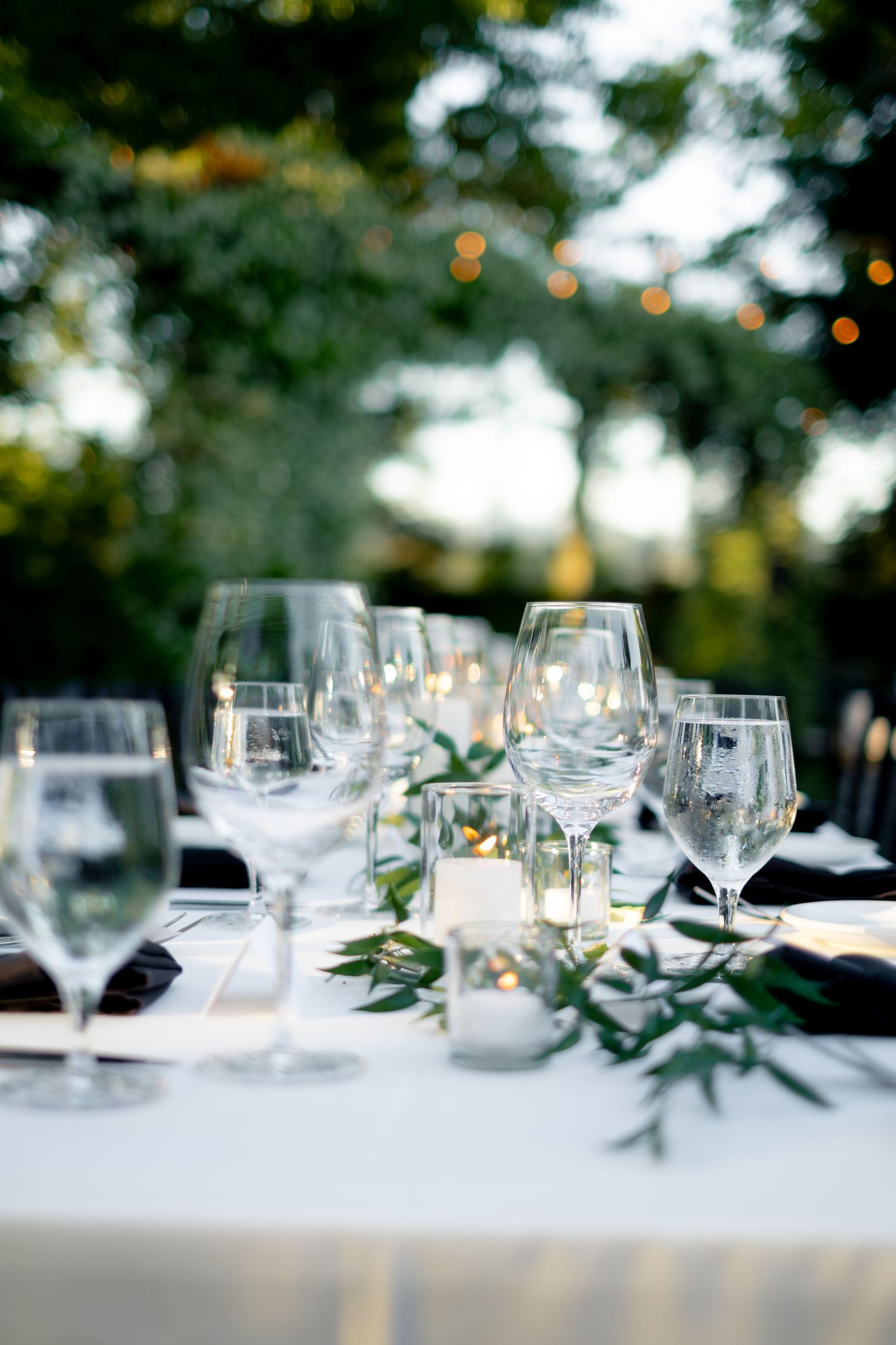 Close-up of crystal wine glasses and greenery centerpieces on a white linen table at an outdoor wedding reception.