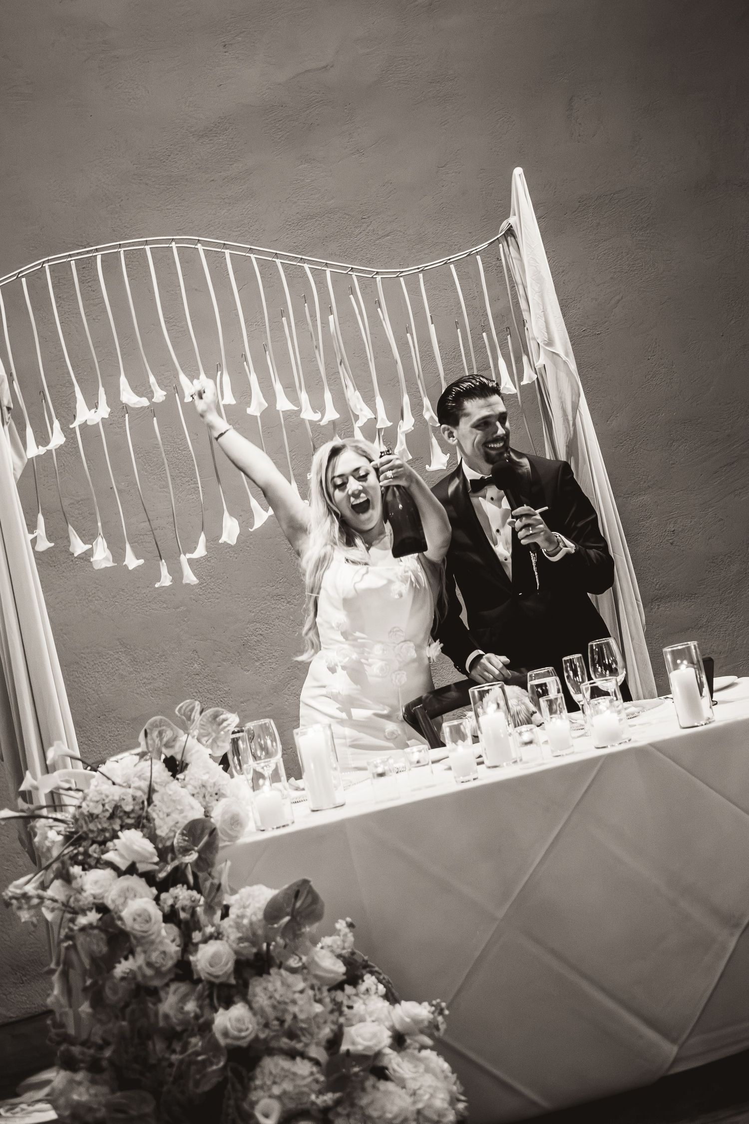Bride and groom celebrate joyfully at wedding reception head table with champagne in black and white photo.