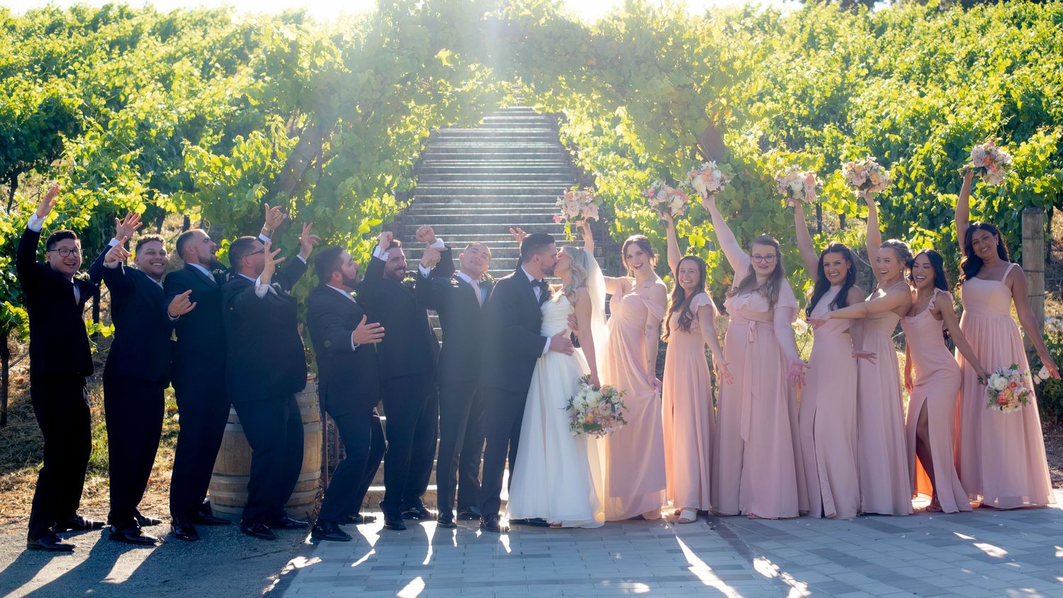 Wedding party celebrates under a vine-covered archway with bridesmaids in blush pink dresses.