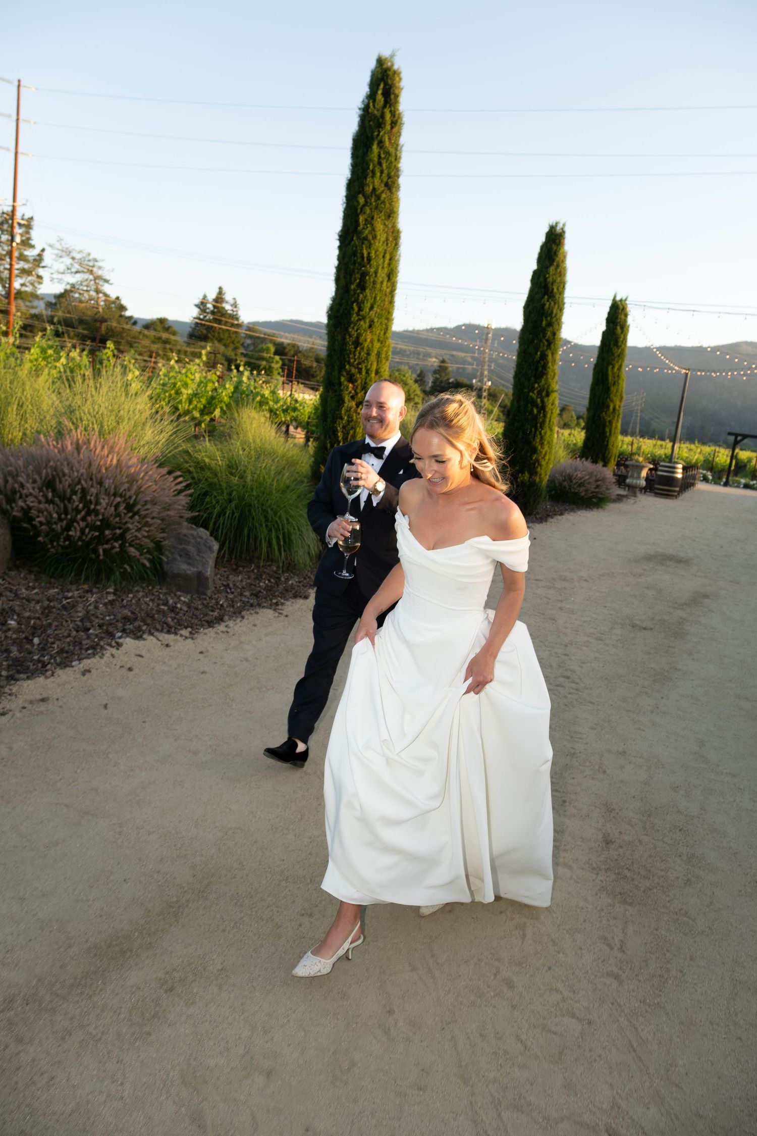 Bride in off-shoulder white gown and groom with champagne strolling along a winery path at dusk.