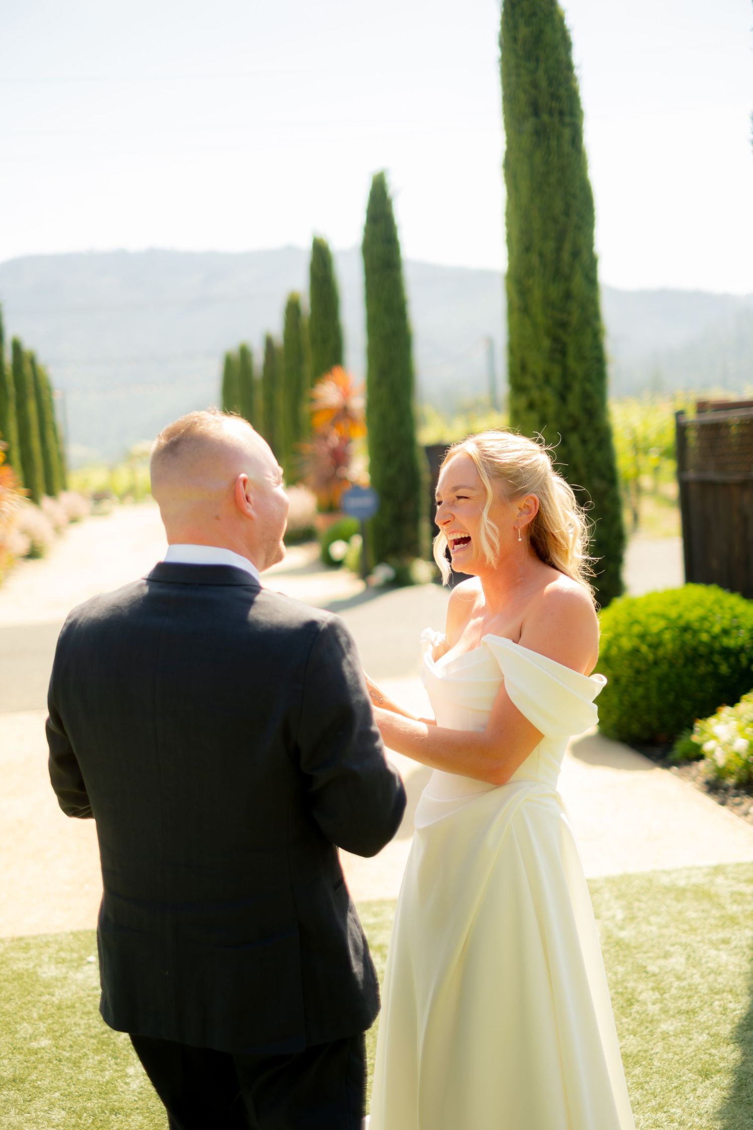 Bride in off-shoulder gown laughing joyfully during first look with groom at vineyard wedding venue.