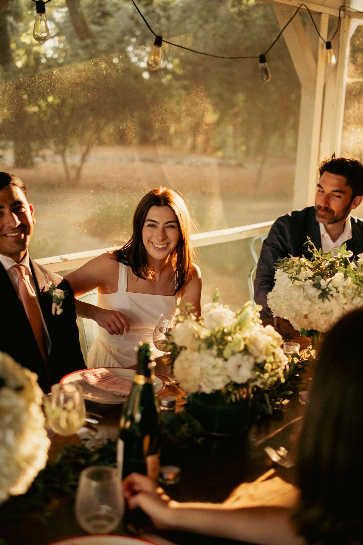 Smiling bride in white dress seated at elegant floral wedding reception dinner table with guests.