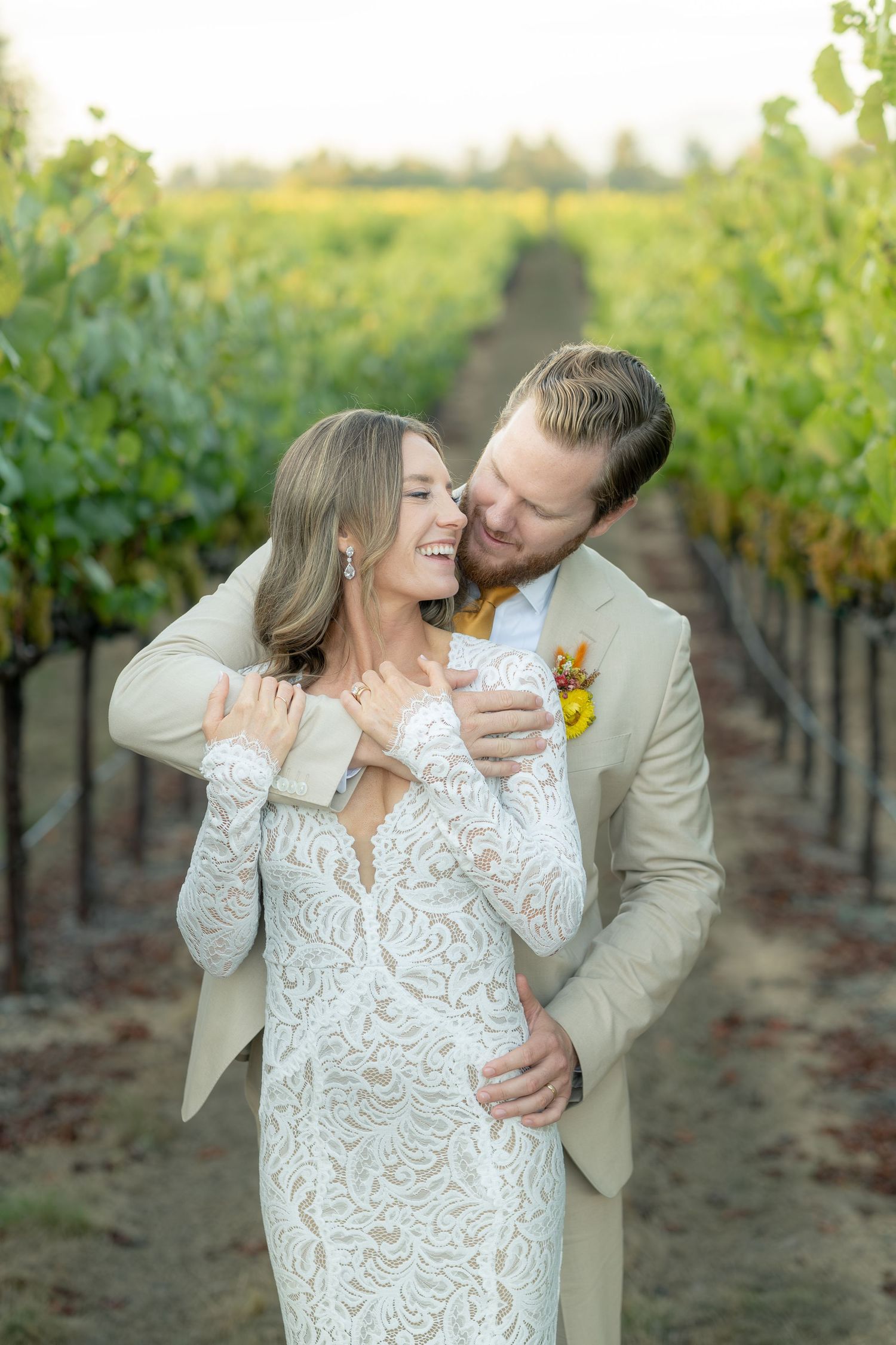 Groom in tan suit embraces laughing bride in lace wedding dress between rows of vineyard grapevines.
