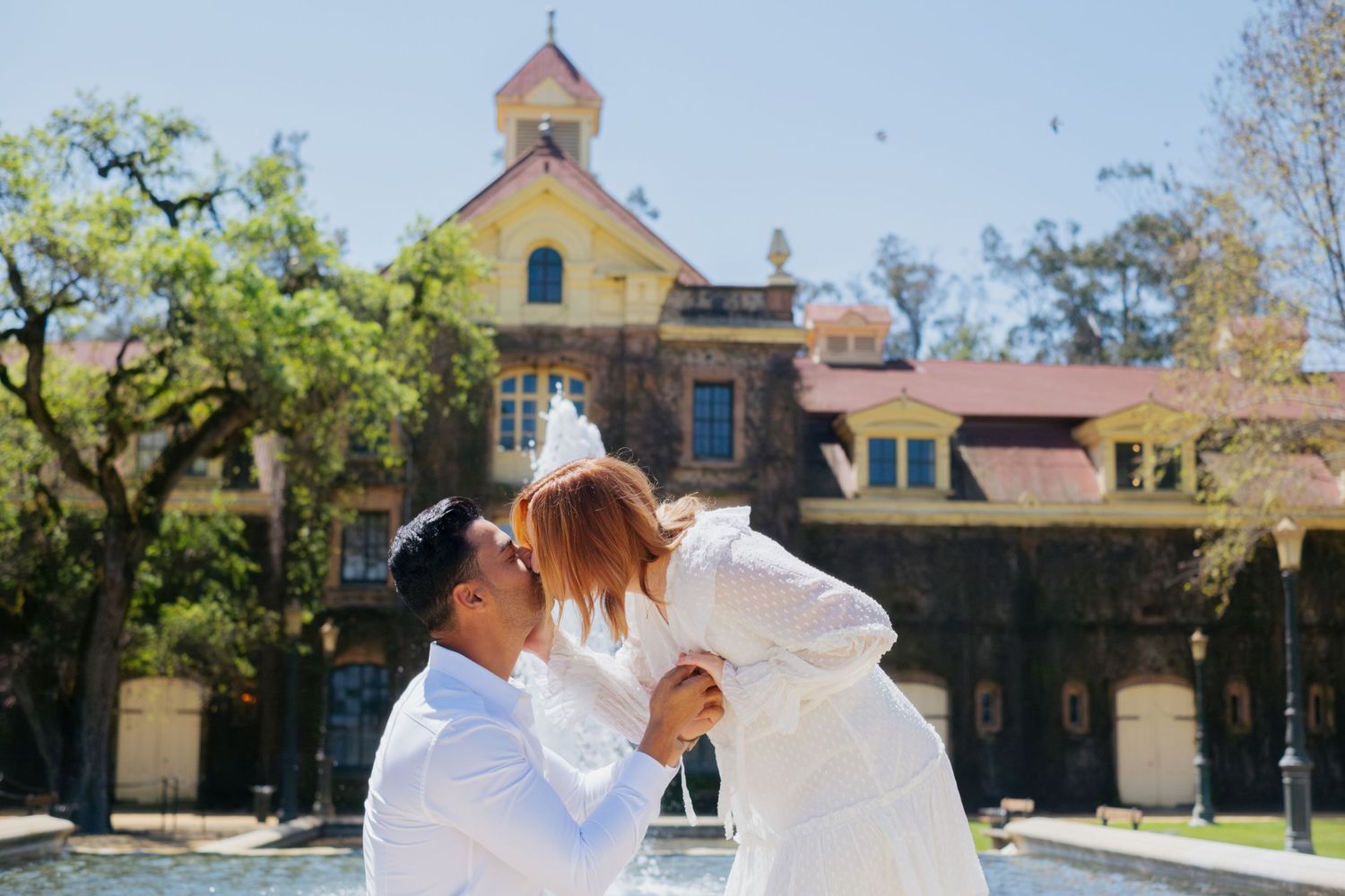 Couple sharing a romantic kiss outdoors in front of a historic yellow building on a sunny day.