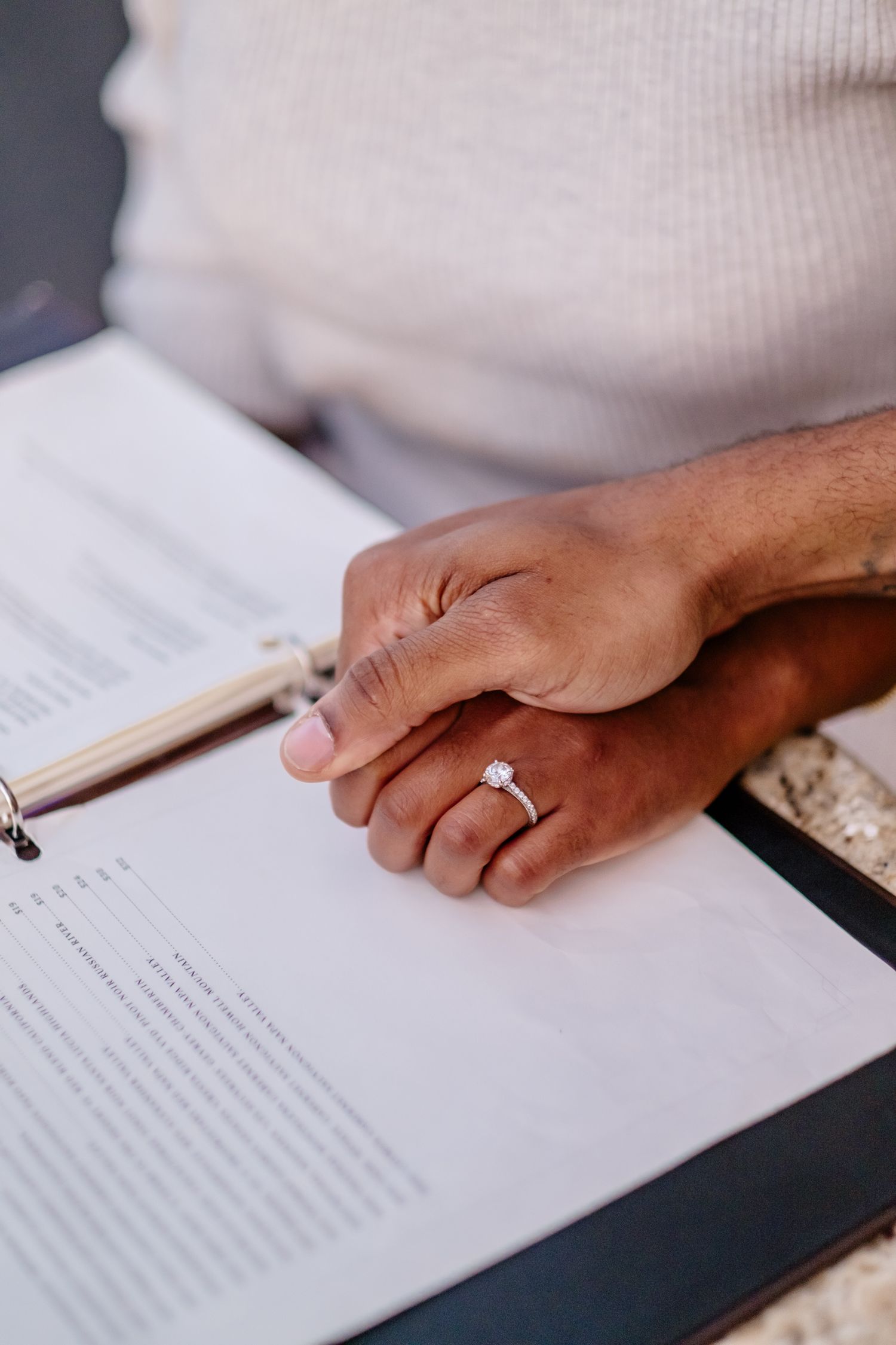 A close-up of two hands resting together on an open book, one adorned with a diamond engagement ring.