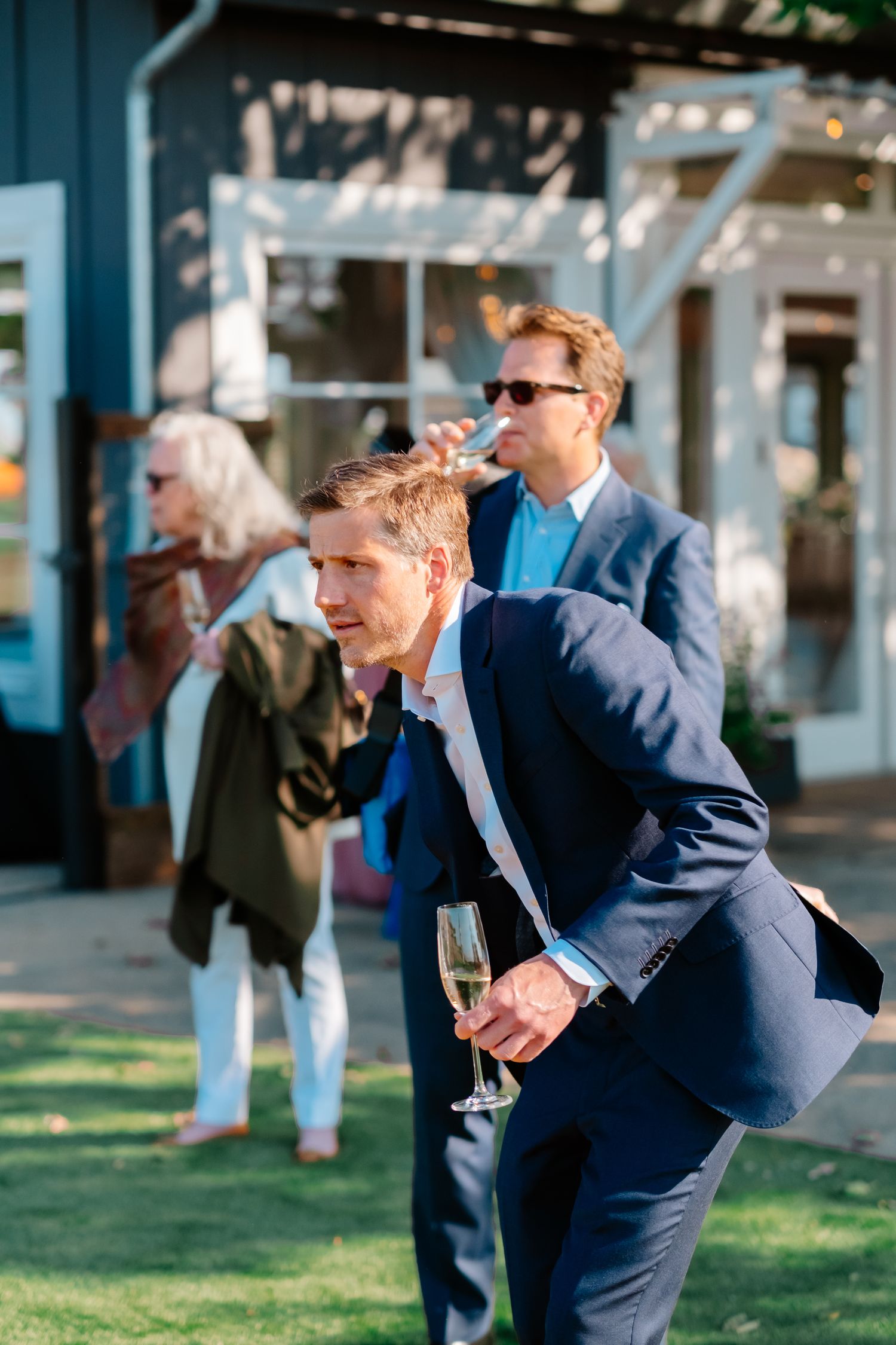 Wedding guests in formal attire enjoy champagne outdoors near a blue building during cocktail hour.