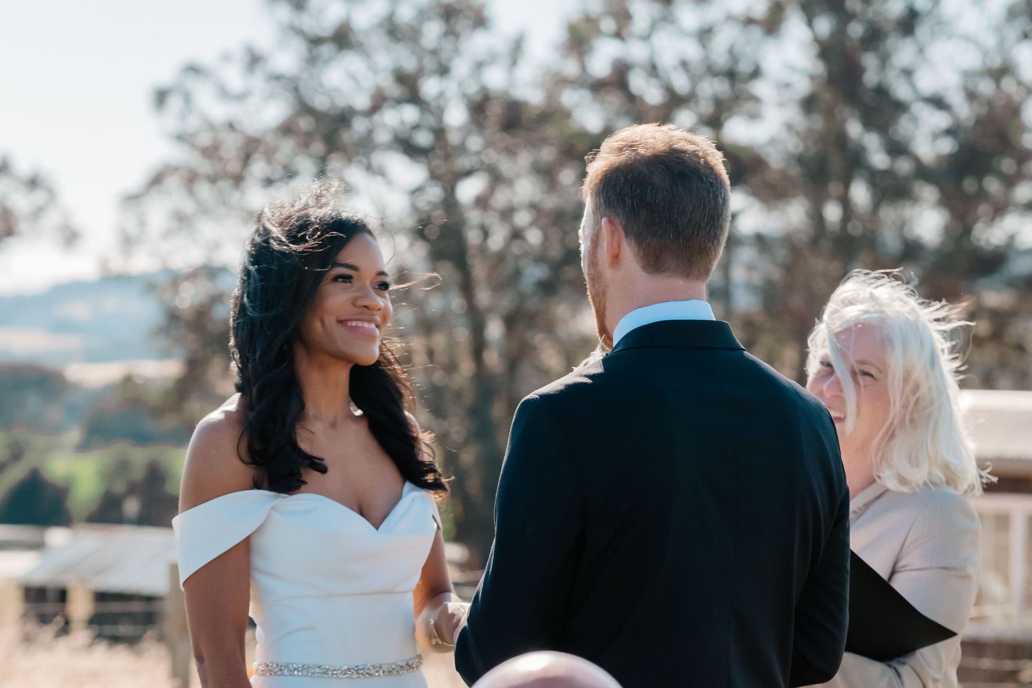 Smiling bride in off-shoulder gown faces groom during outdoor wedding ceremony.