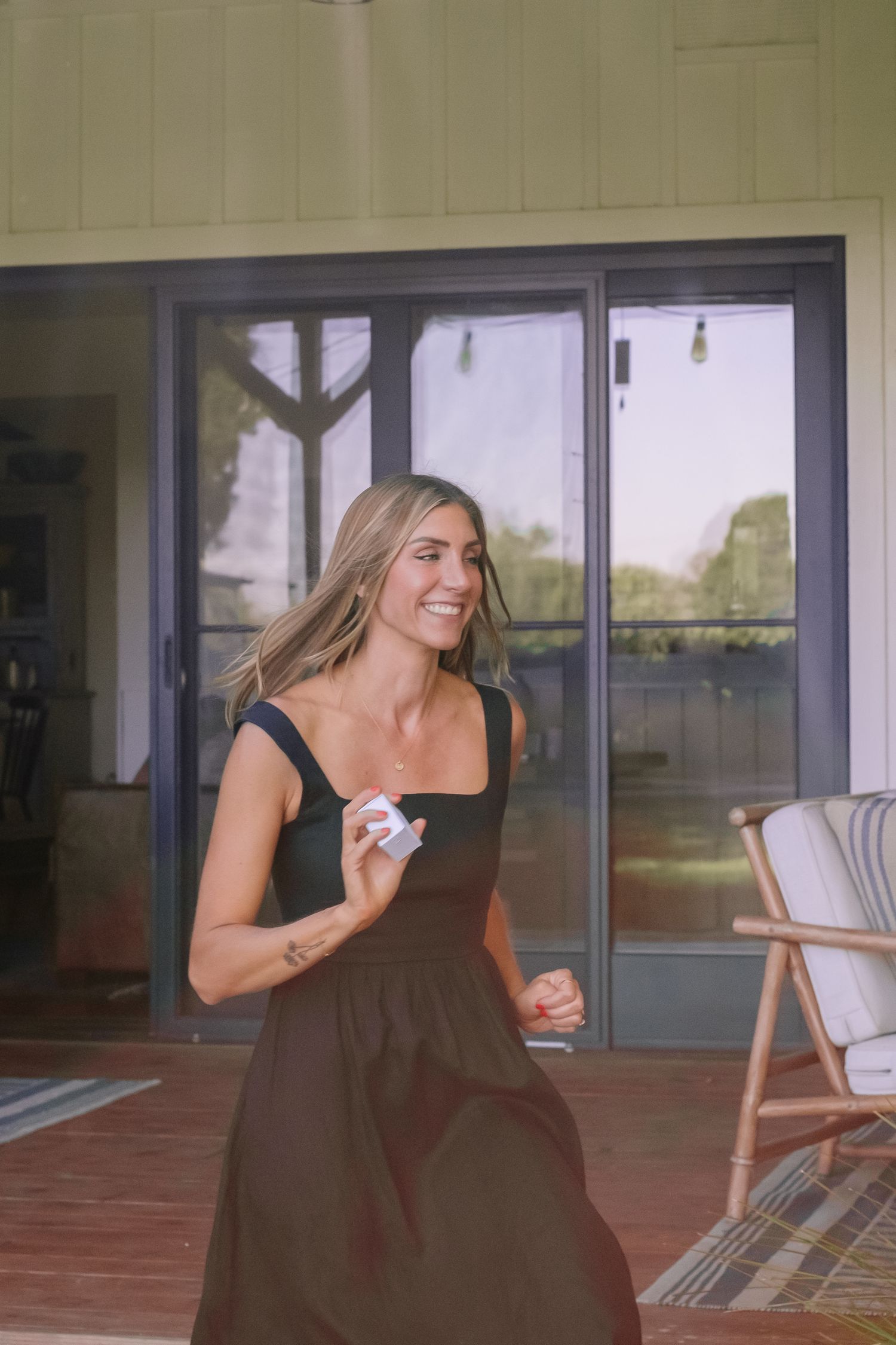 Smiling blonde woman in black dress walks on wooden deck outside modern green building.