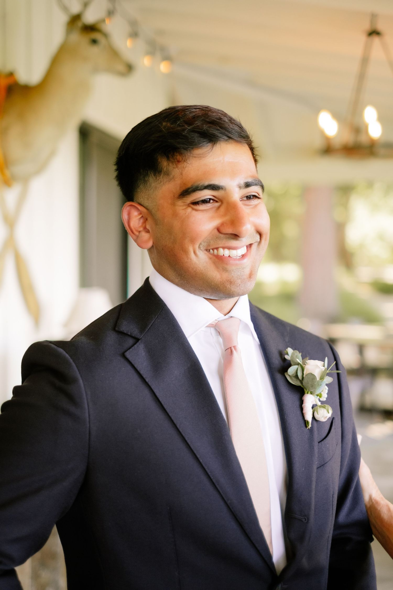 Smiling groom in dark suit with blush tie and floral boutonniere on wedding day.