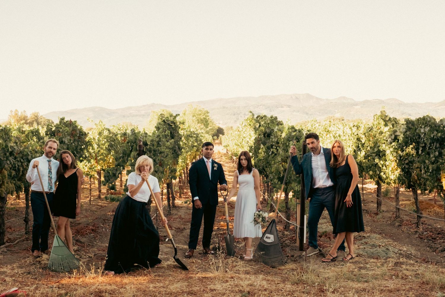 Wedding party poses with garden tools in vineyard at sunset with mountain backdrop.