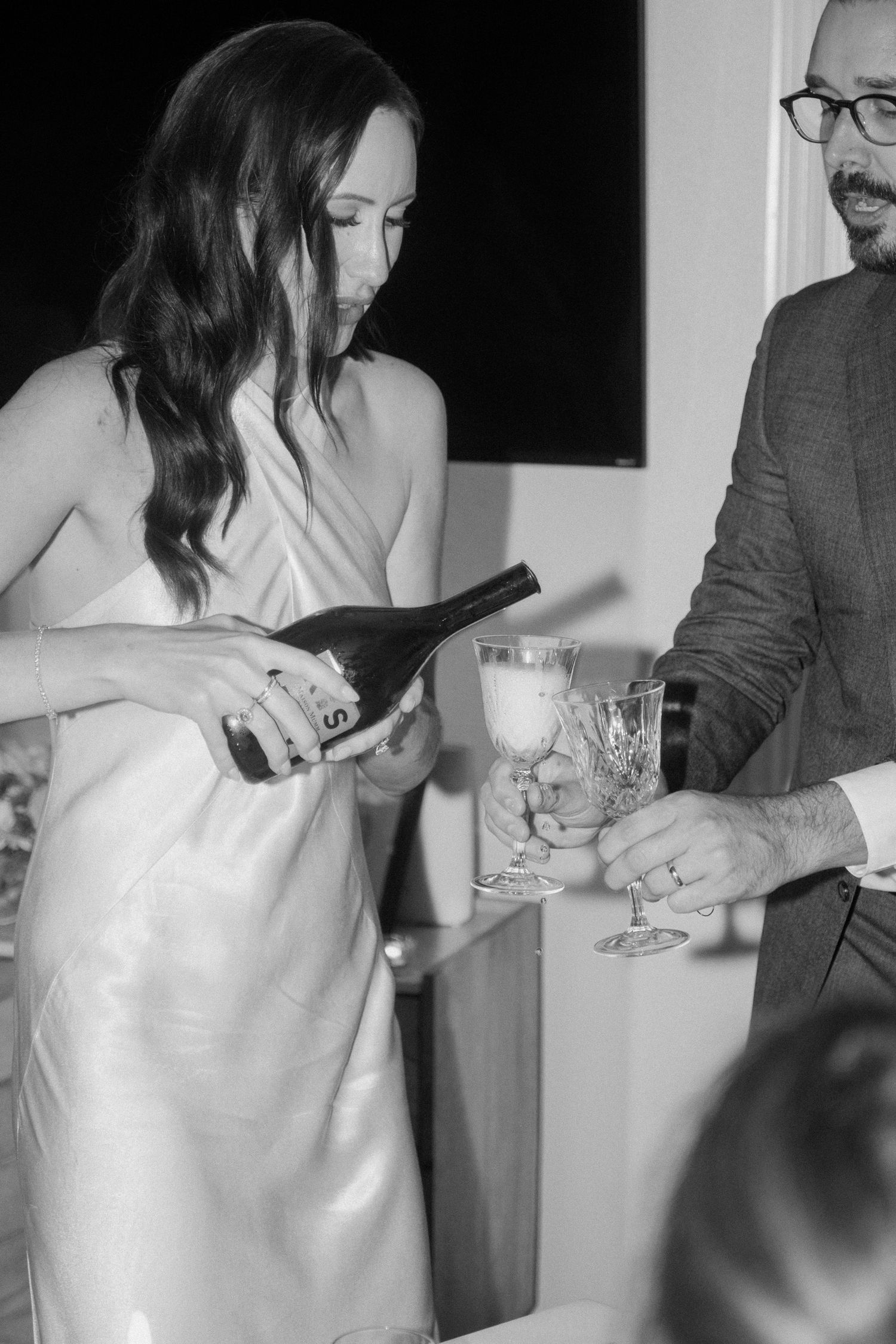 Bride in white slip dress pouring champagne into crystal glasses at wedding reception in black and white photo.