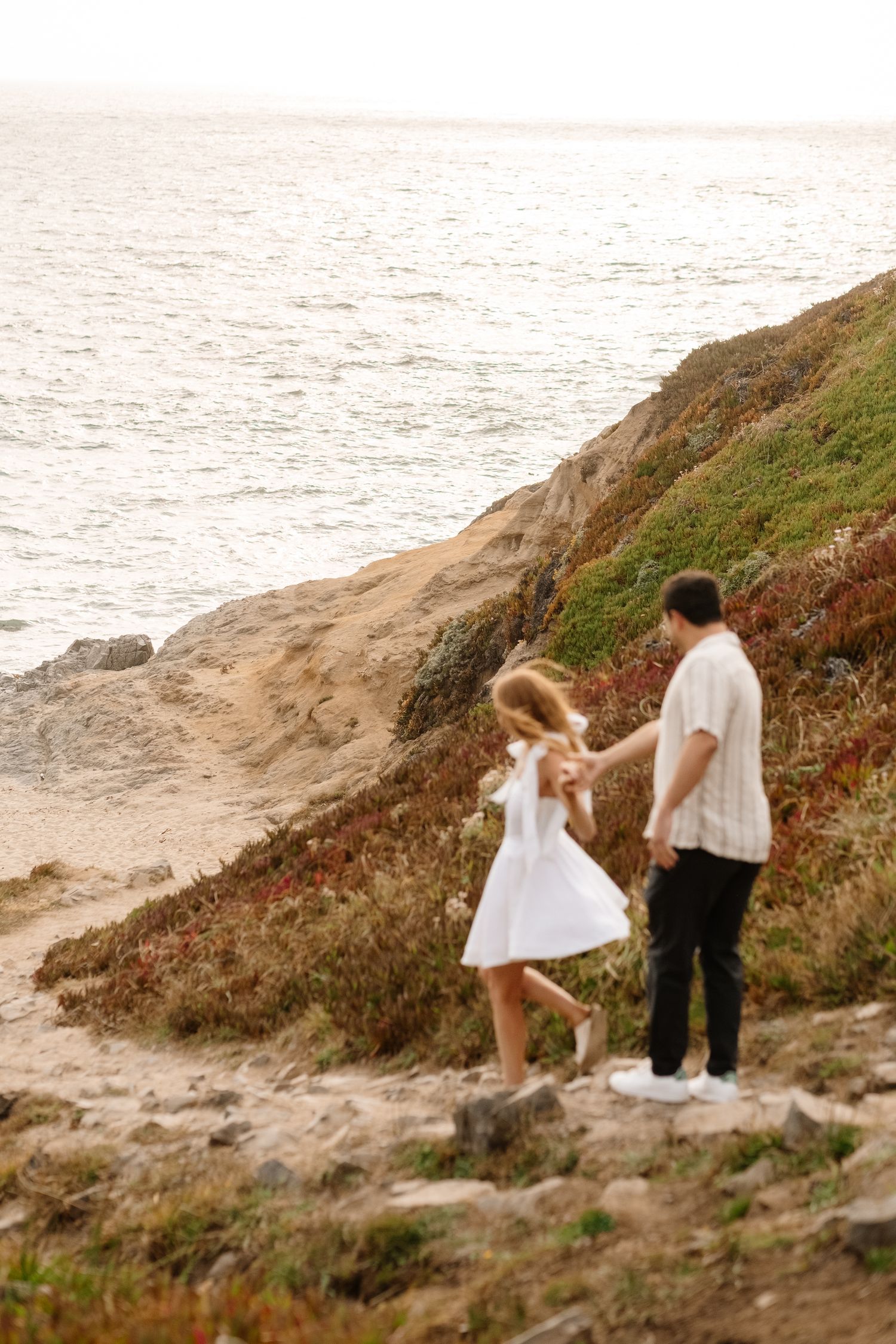 A couple holds hands while walking along a scenic coastal cliffside path overlooking the ocean.