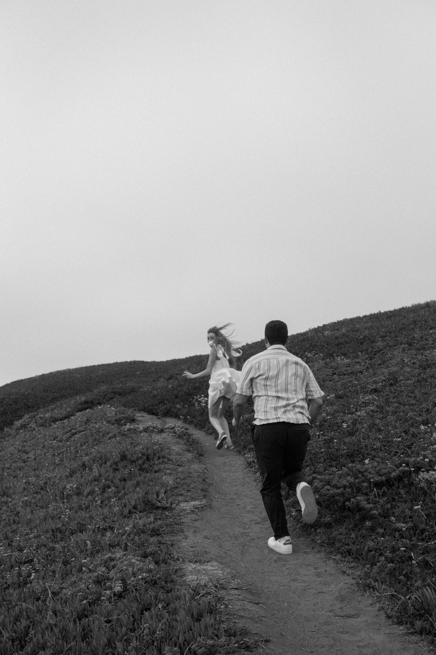 A couple runs up a grassy hillside path together in this black and white outdoor photo.