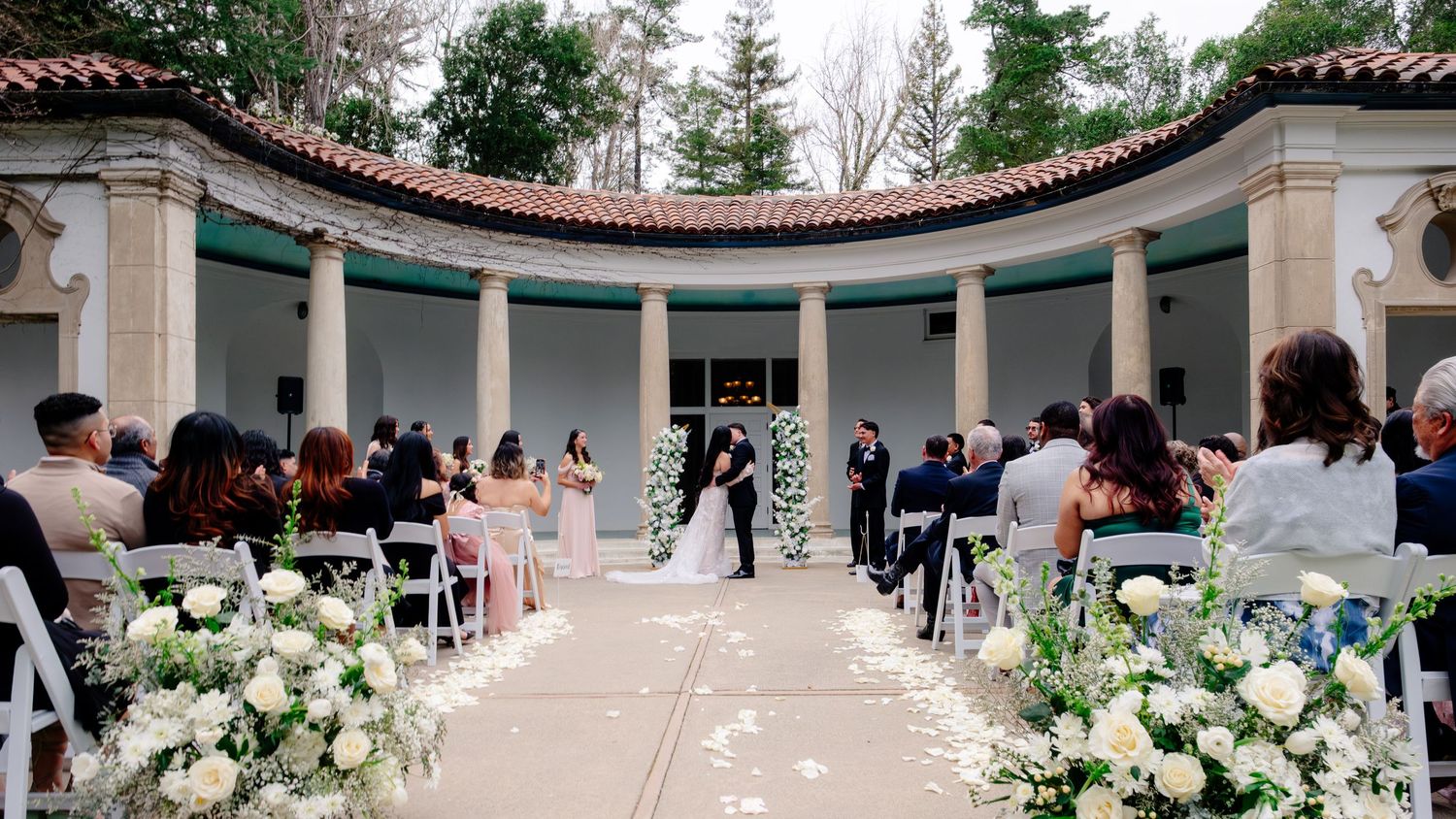 Elegant outdoor wedding ceremony at a classical pavilion with white floral arrangements lining the aisle.