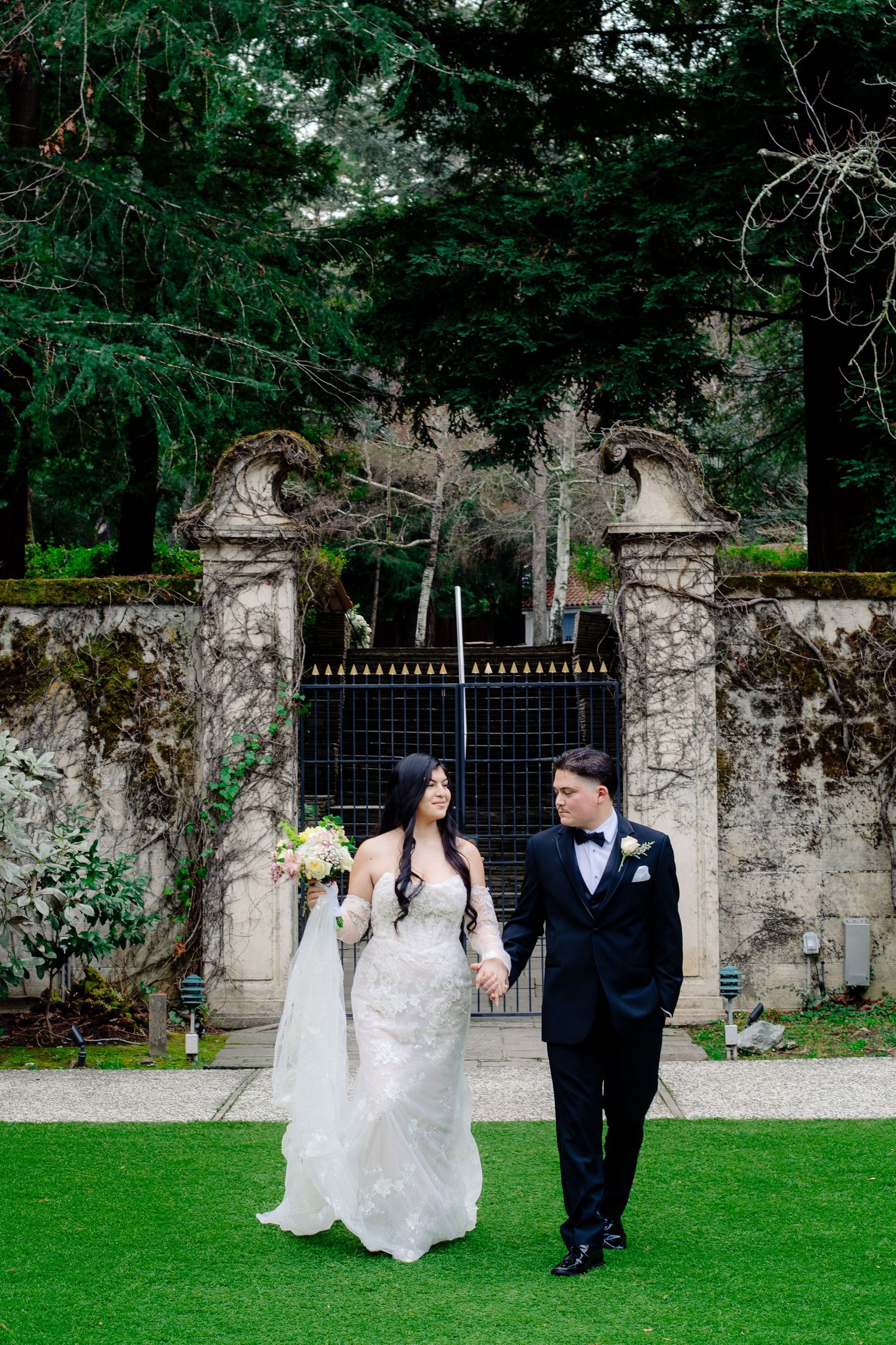 Bride and groom walking hand in hand on green lawn before stone gate and tall evergreen trees.