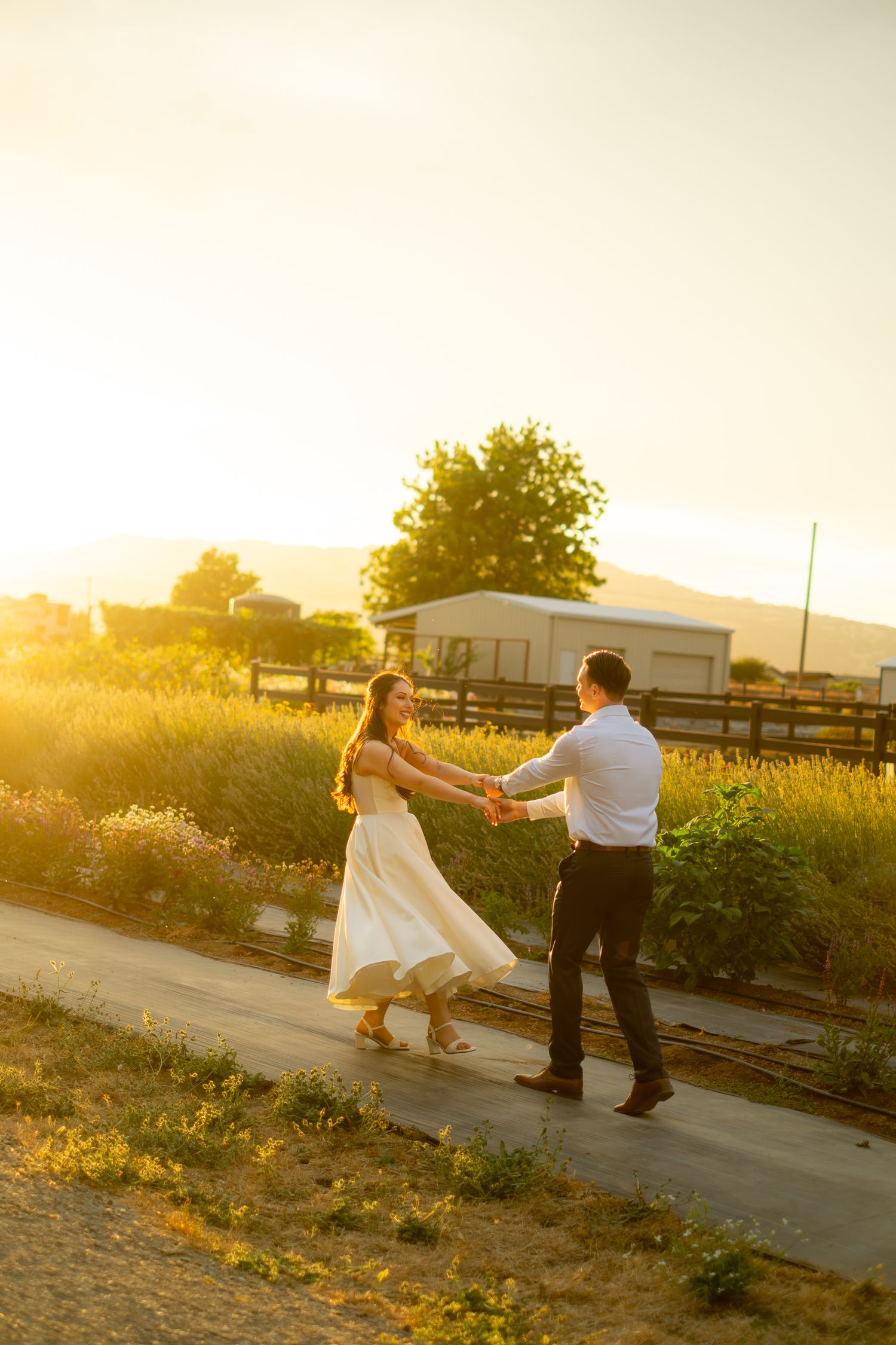 Newlyweds dance joyfully on a sunlit path surrounded by lavender fields at sunset.