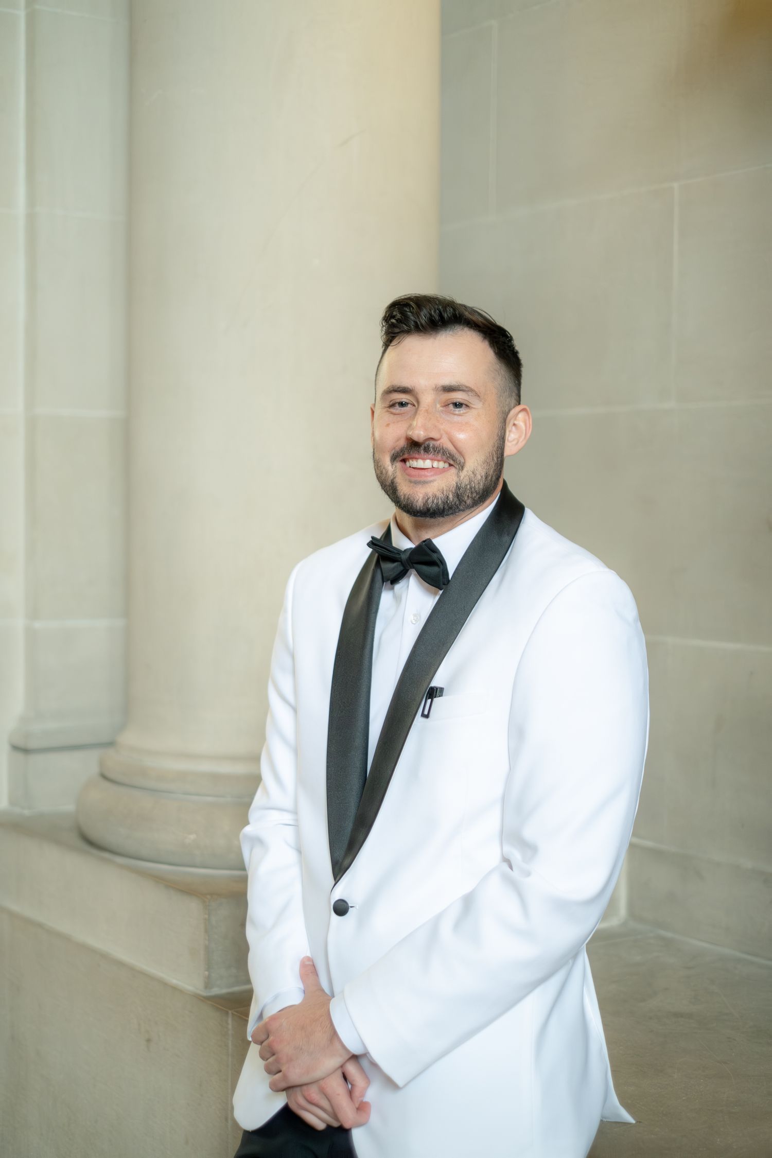 Smiling groom in white tuxedo jacket with black lapels and bow tie standing by stone columns.