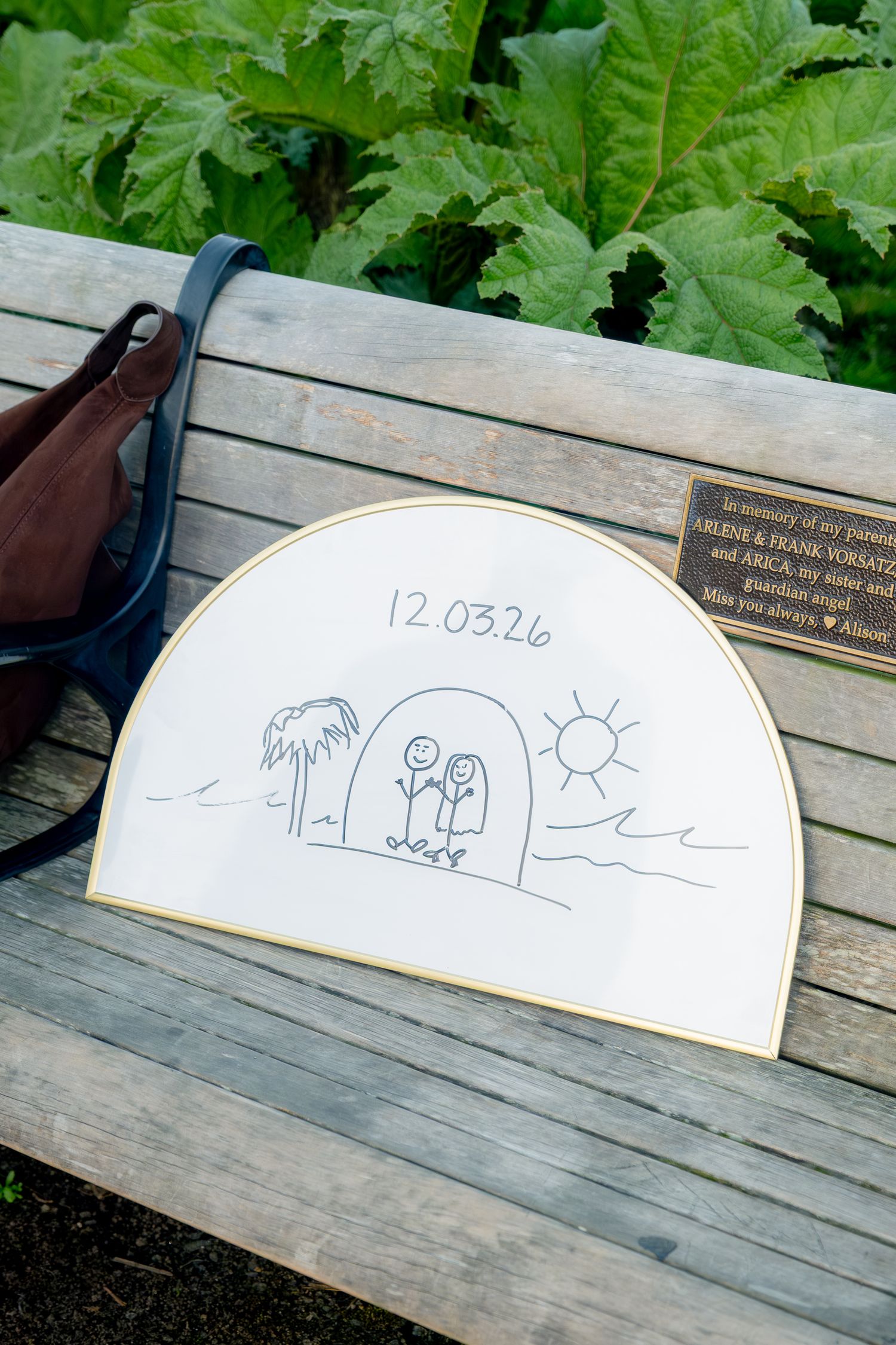 A semicircular drawing dated 12.03.26 with stick figures rests on a wooden memorial bench beside a leather bag.