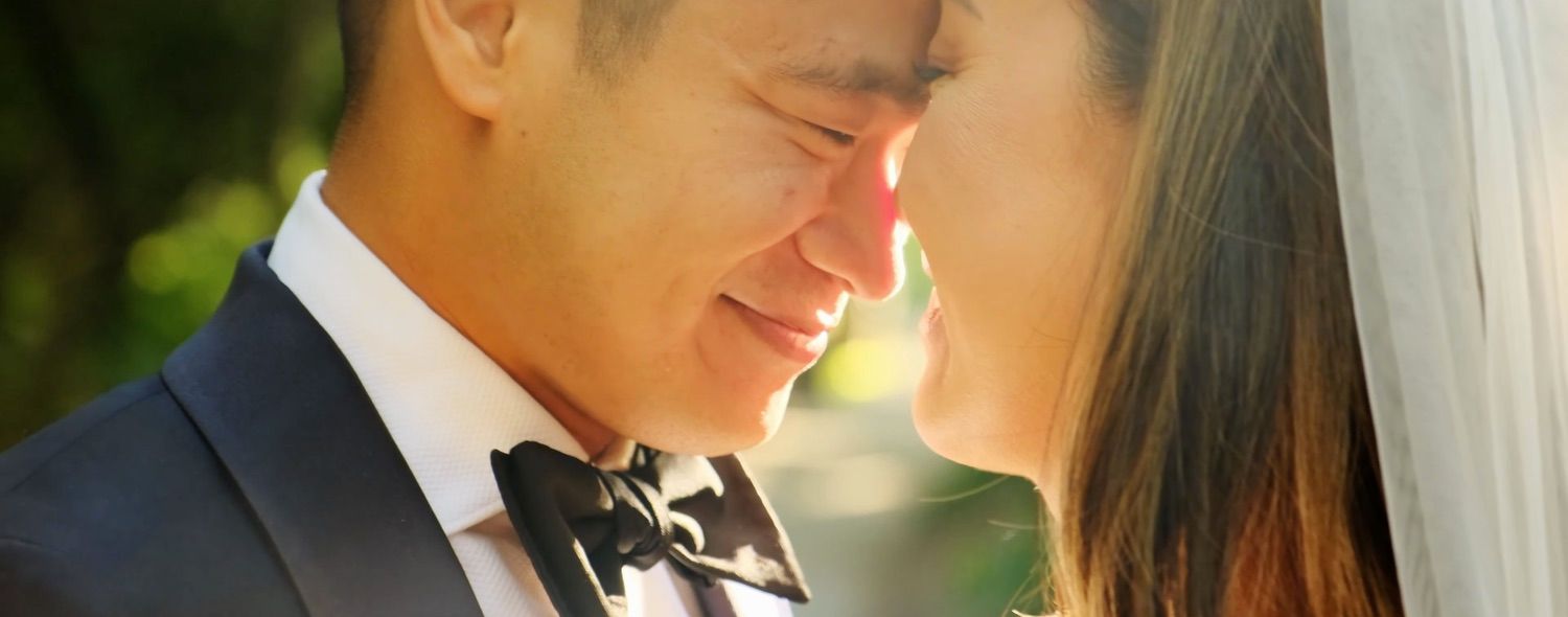 Close-up of smiling groom in bow tie sharing a tender forehead touch with bride in sunlight.