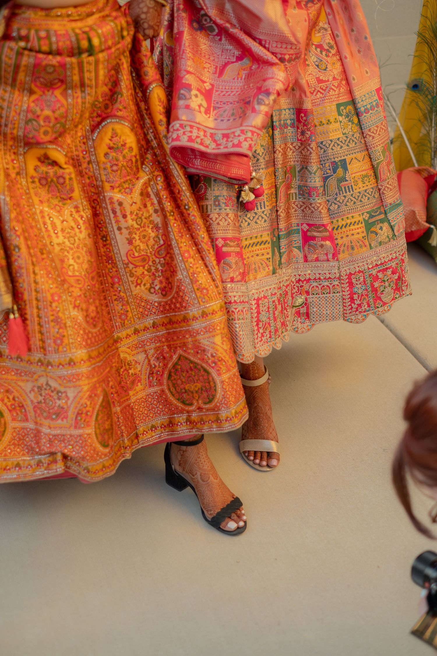 Close-up of vibrant orange and pink embroidered lehenga skirts with sandals at Indian wedding.