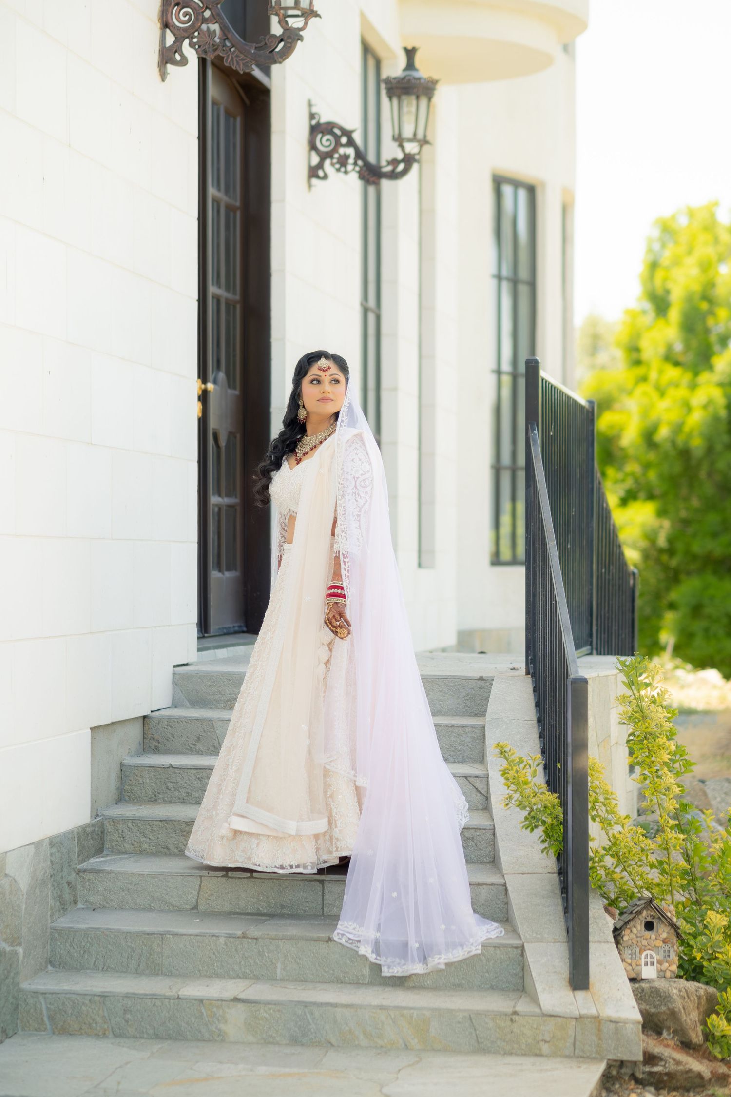 Indian bride in white lehenga and veil stands gracefully on stone steps outside elegant white venue.