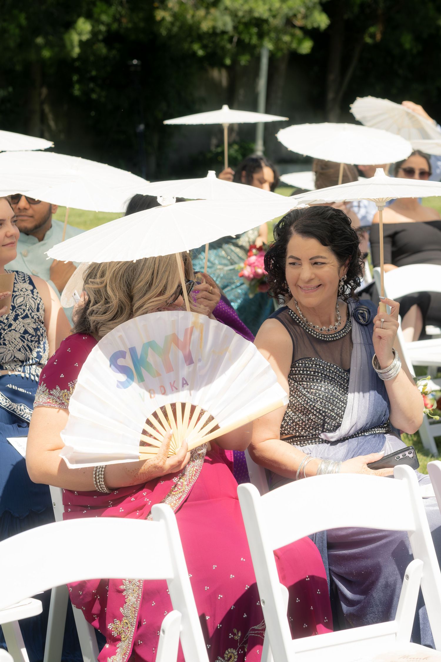 Wedding guests use white parasols and fans to shade from sun at outdoor ceremony.