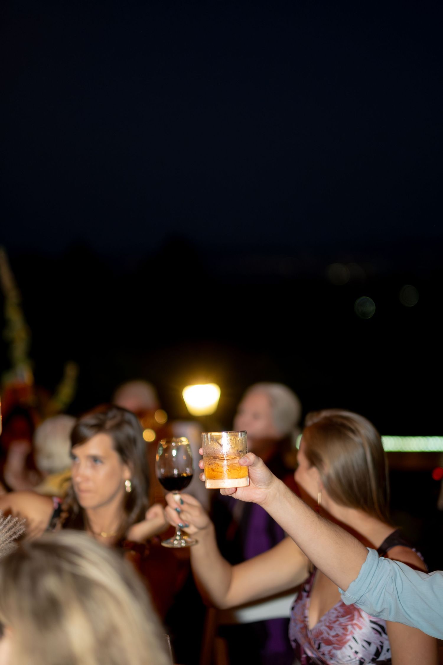 Wedding guests raising glasses for a toast at night, featuring whiskey and red wine glasses outdoors.