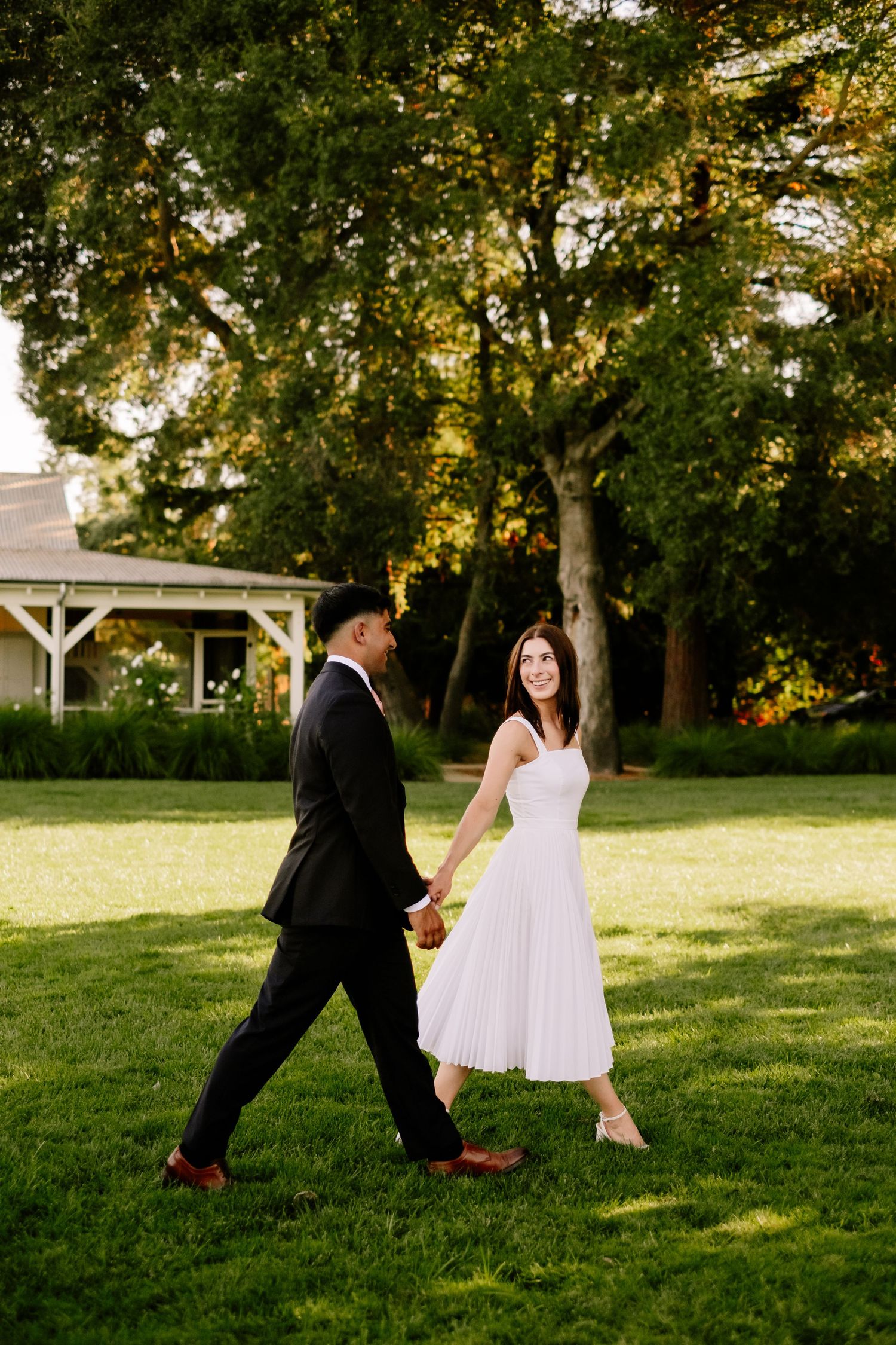Bride and groom holding hands walking across lush green lawn at golden hour.