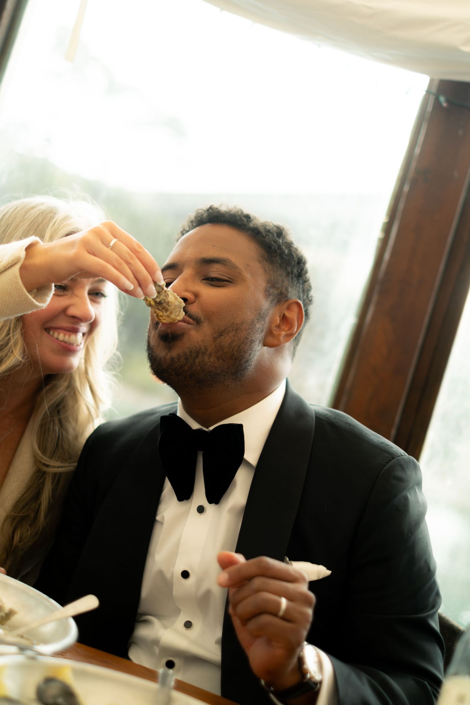 Groom in tuxedo eating oyster fed by smiling blonde bride at wedding reception dinner.